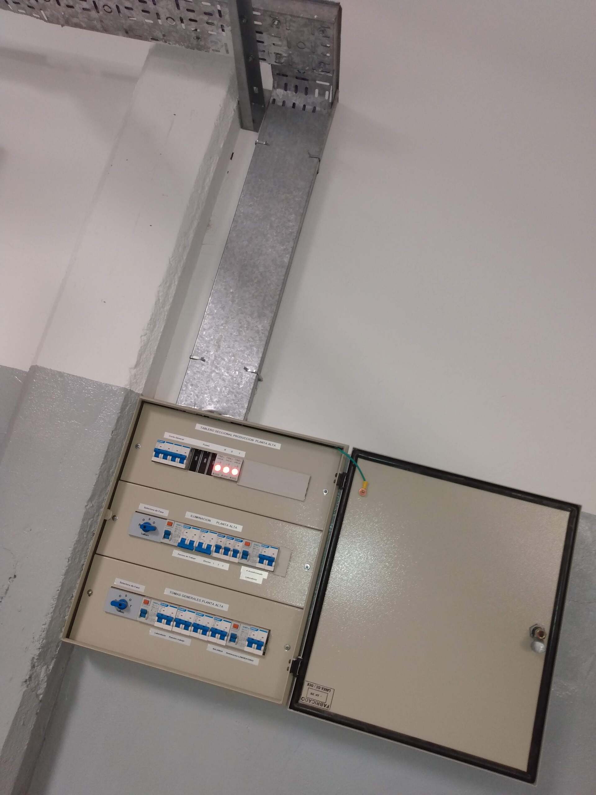 Una caja de interruptores eléctricos abierta montada en la pared con tres filas de interruptores, ubicada debajo de una bandeja metálica portacables.