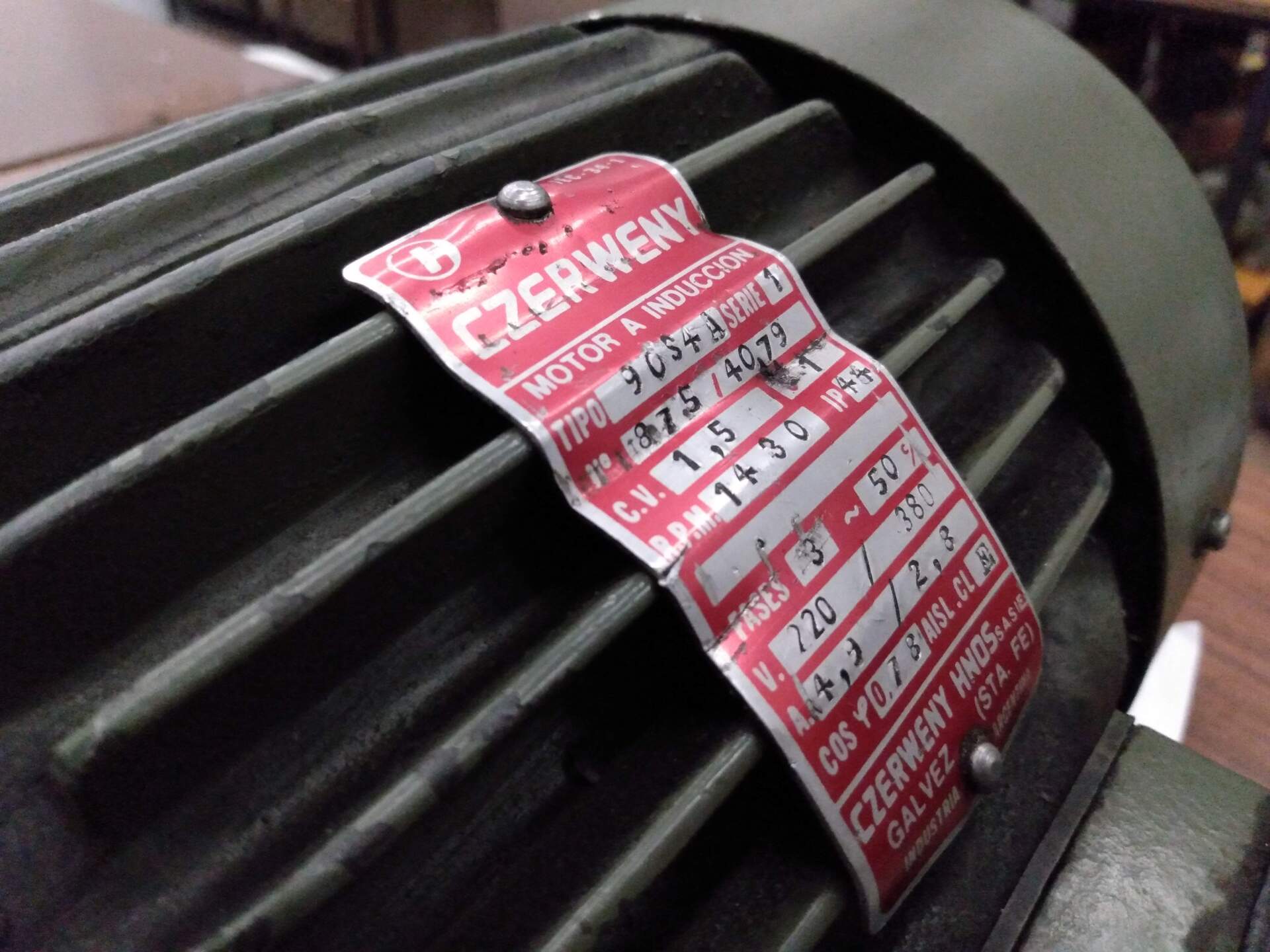 Una placa roja con el nombre del motor, fijada a un motor eléctrico verde acanalado, que muestra las especificaciones técnicas y los datos de la marca.