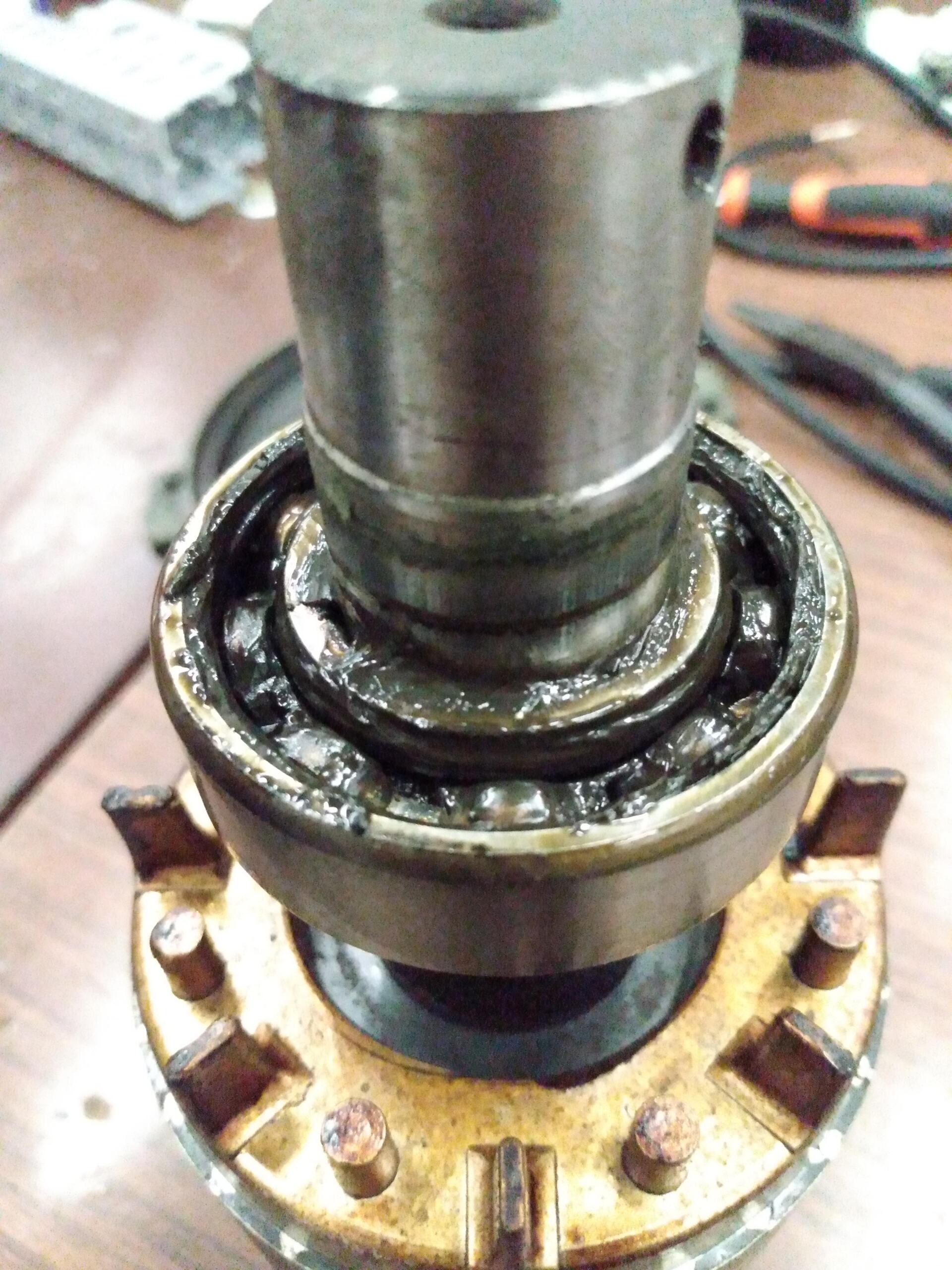 Primer plano de un eje de motor metálico con un conjunto de rodamientos de bolas desgastados y cubiertos de grasa que descansa sobre un componente de latón.