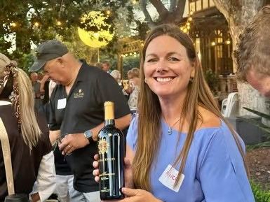 Woman in blue shirt holding a wine bottle and smiling outdoors at a gathering