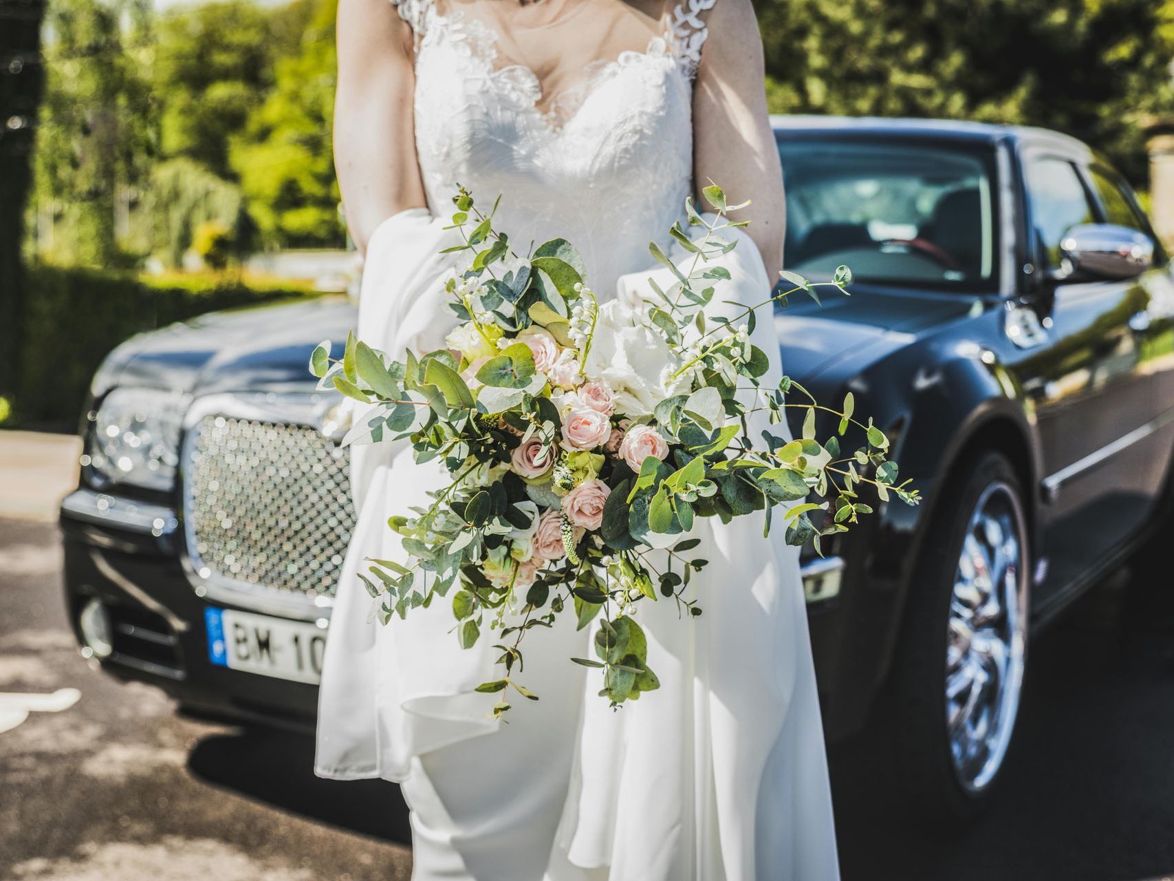 A bride in a wedding dress is holding a bouquet of flowers in front of a black car.