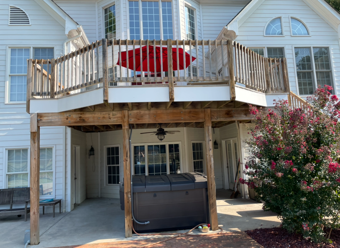Two-story white house with a deck; red umbrella visible. Hot tub beneath the deck, near flowering bush.