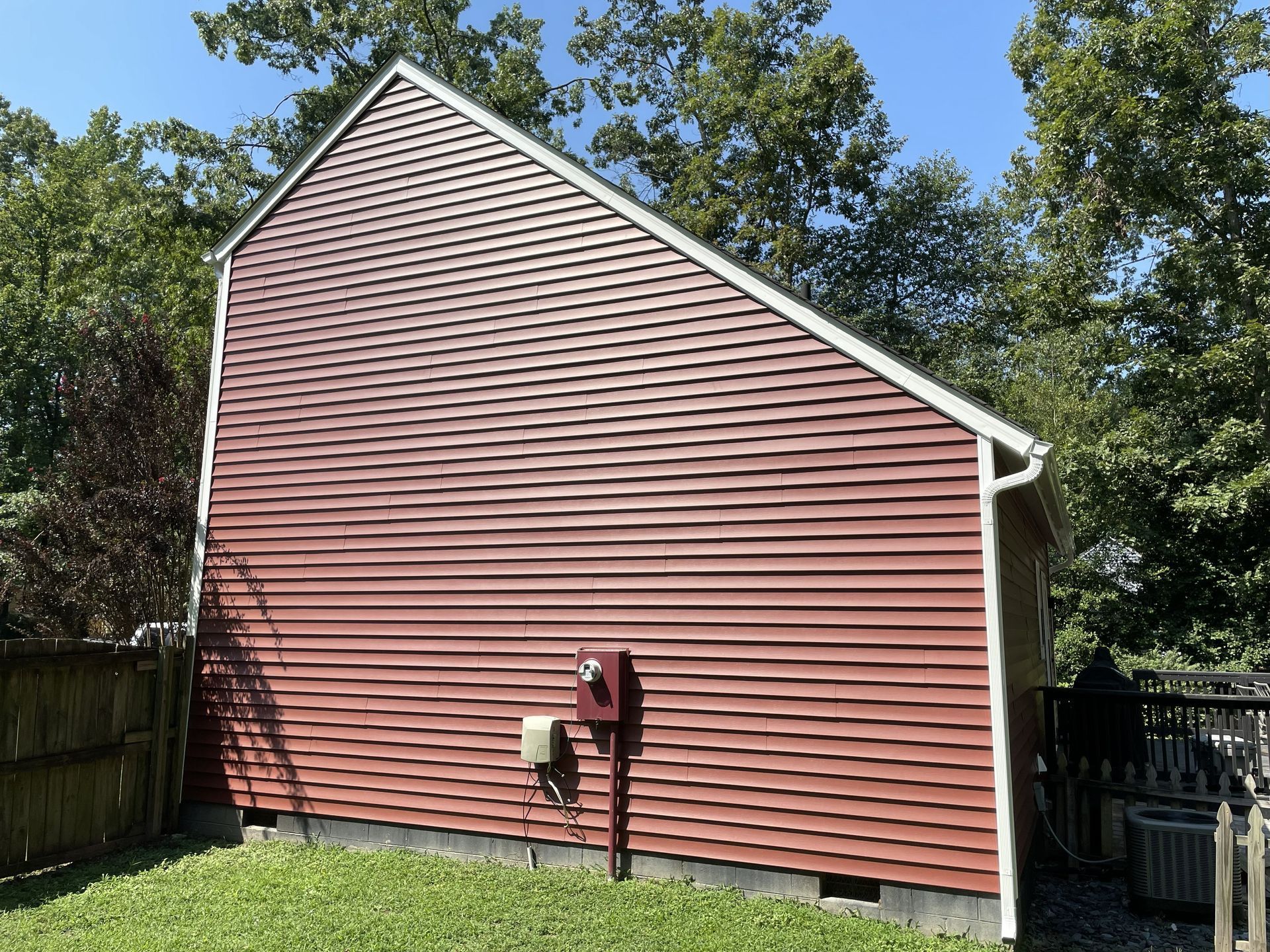 Red siding on a building with white trim; a small electrical box is visible. Green grass and trees are in the background.