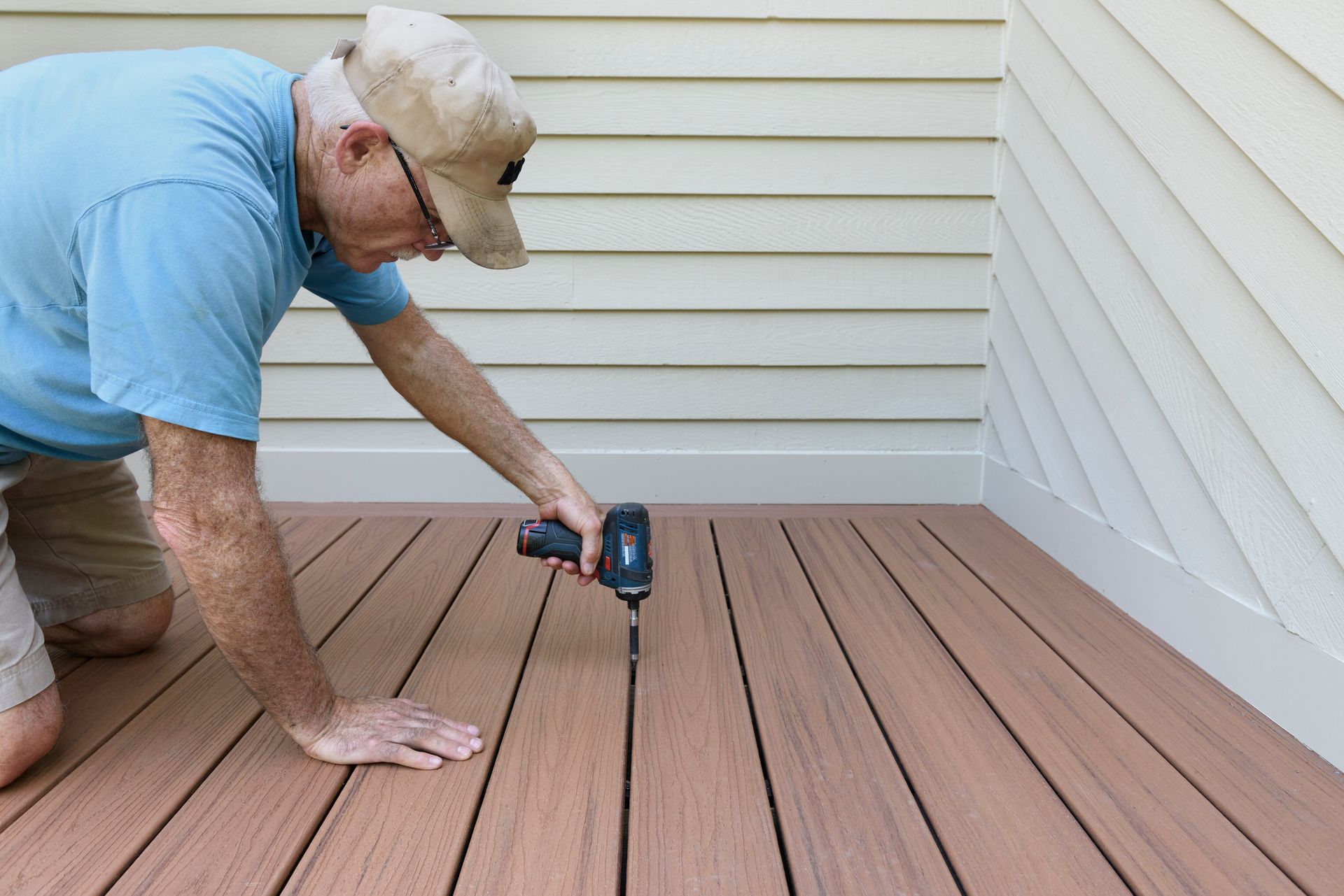 Man kneeling on a wooden deck using a power drill to fasten deck boards near a house exterior.