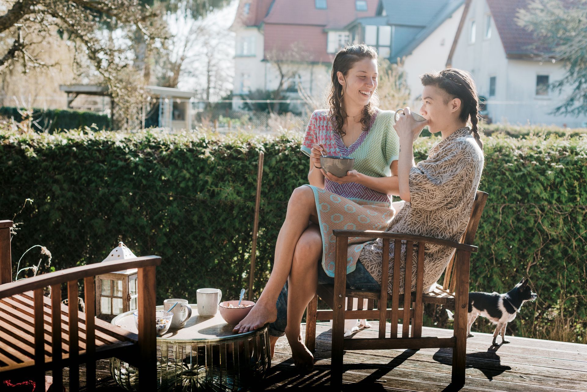 Two people share food on a sunny deck with a small dog nearby.