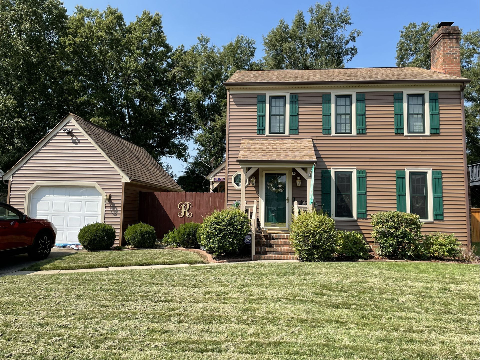 Two-story brown house with green shutters and garage; green lawn and trees in the background.