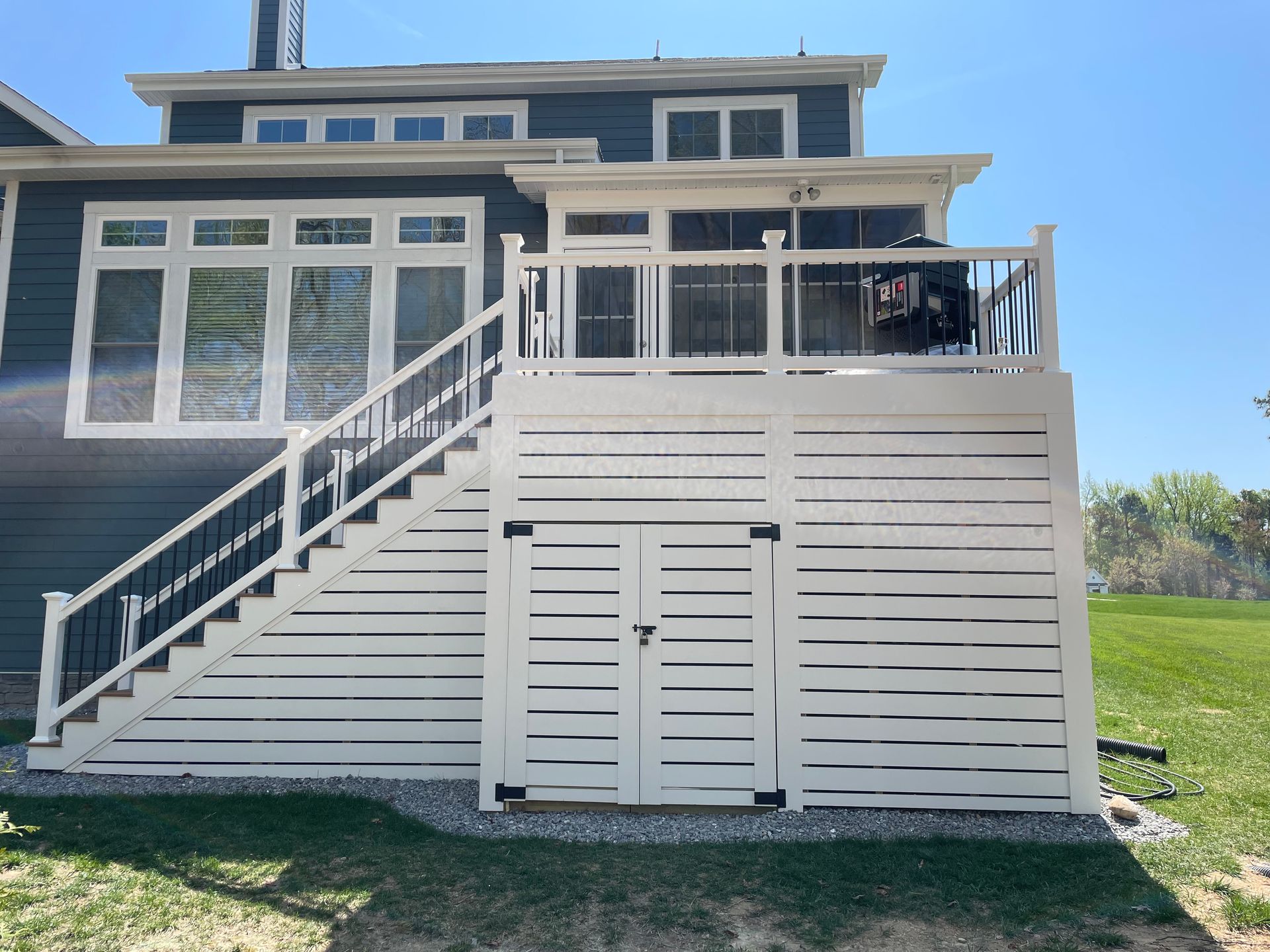 White deck with storage, stairs, and railing against a blue house with windows, sunny day.