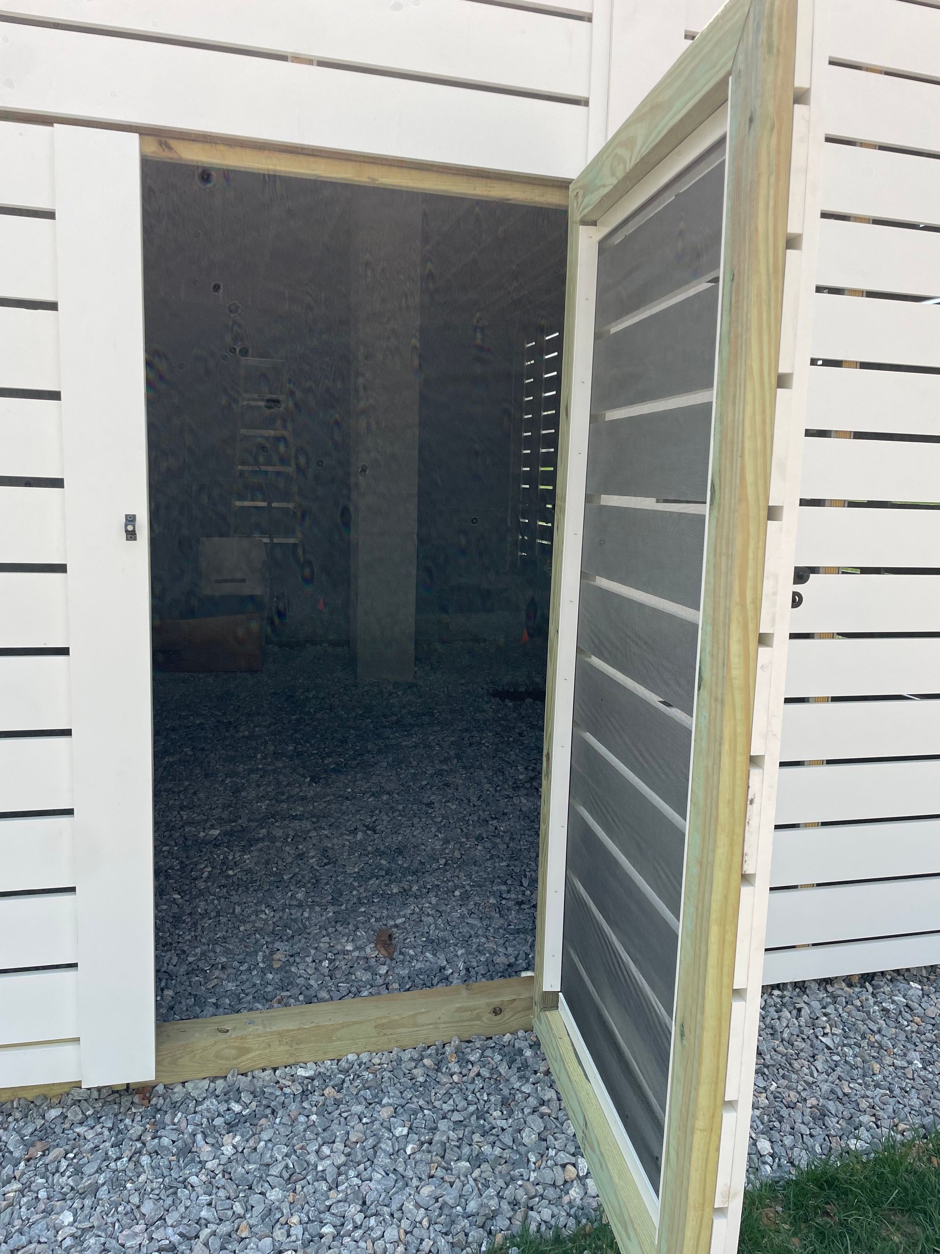 Open door with screen into a gravel-floored crawlspace beneath a white building. Green trim.