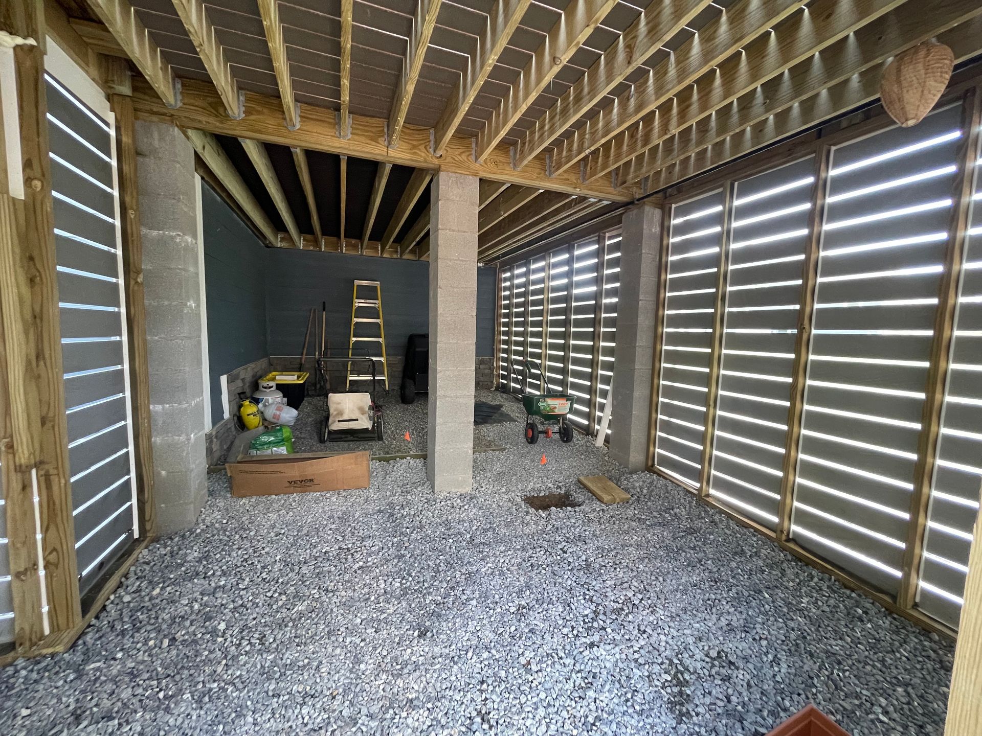 Under-deck storage area with gravel floor, wooden beams, concrete pillars, and slatted walls.