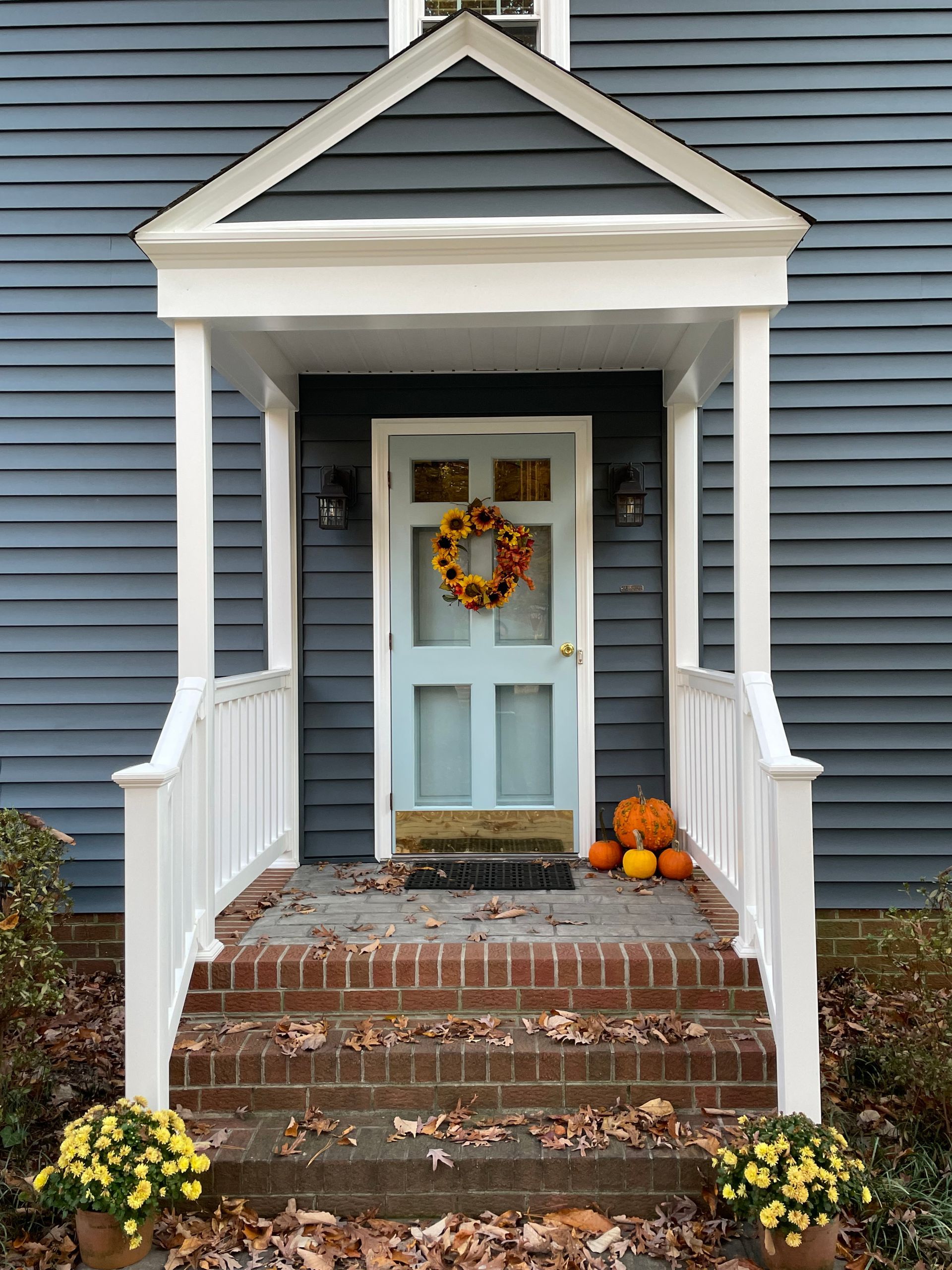 Blue door with fall wreath, porch with white trim, brick steps, pumpkins, and mums.