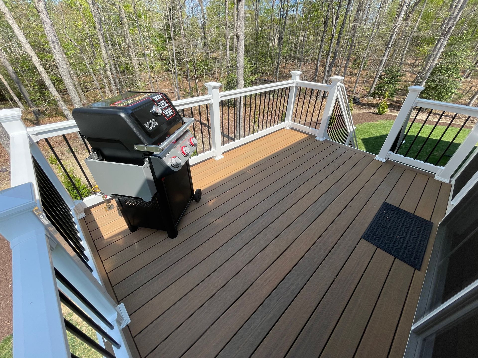 Wooden deck with grill, black railing and white posts, surrounded by trees.