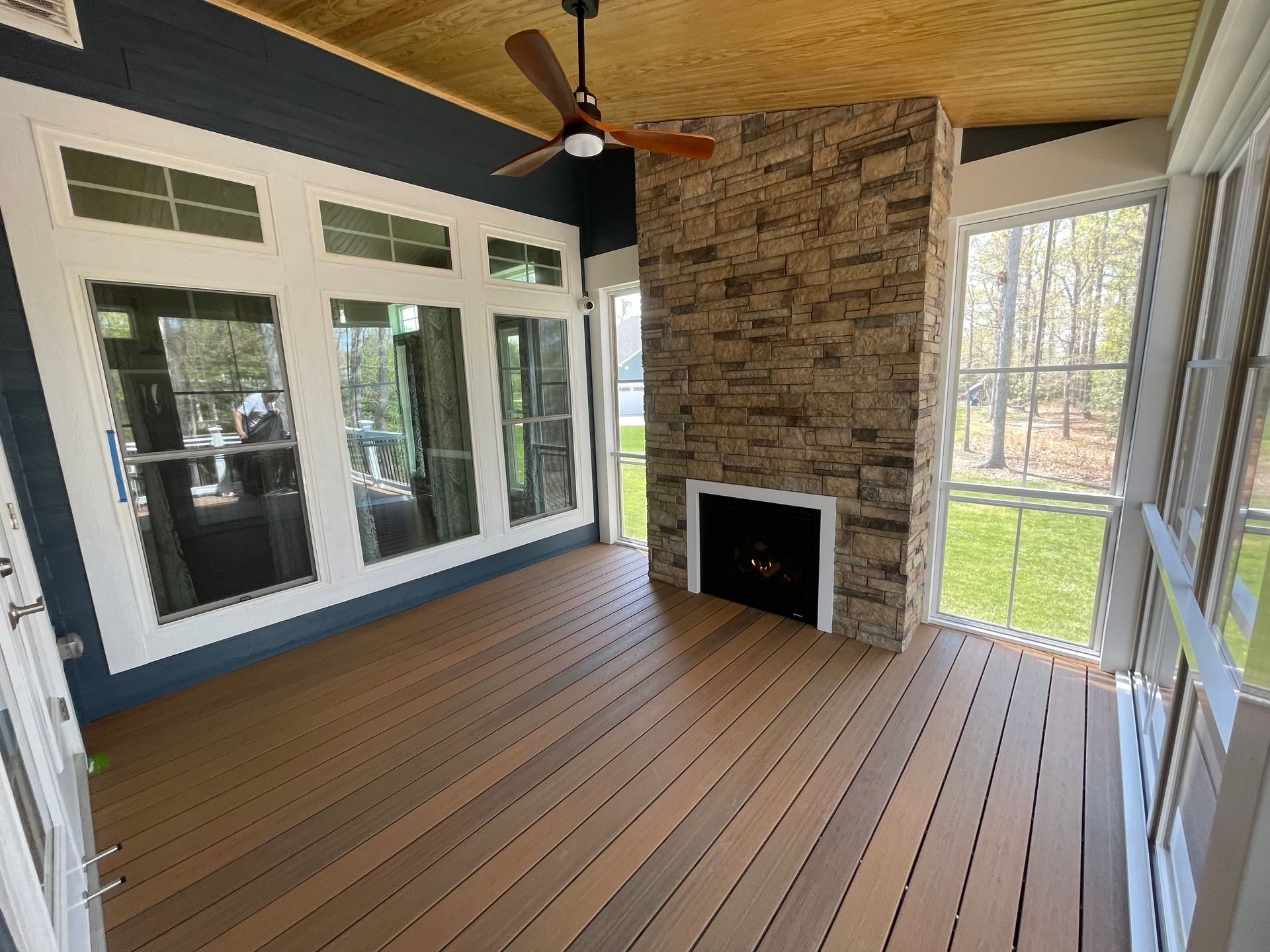 Sunroom with a stone fireplace, composite wood flooring, and large windows overlooking trees.