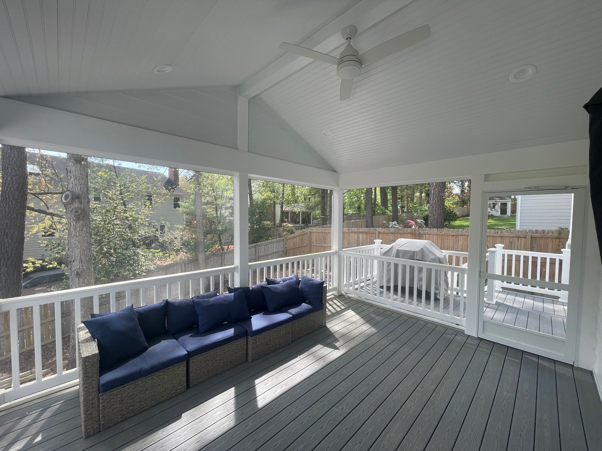 Screened-in porch with blue sofa, white railing, and grey deck, overlooking a backyard with trees.