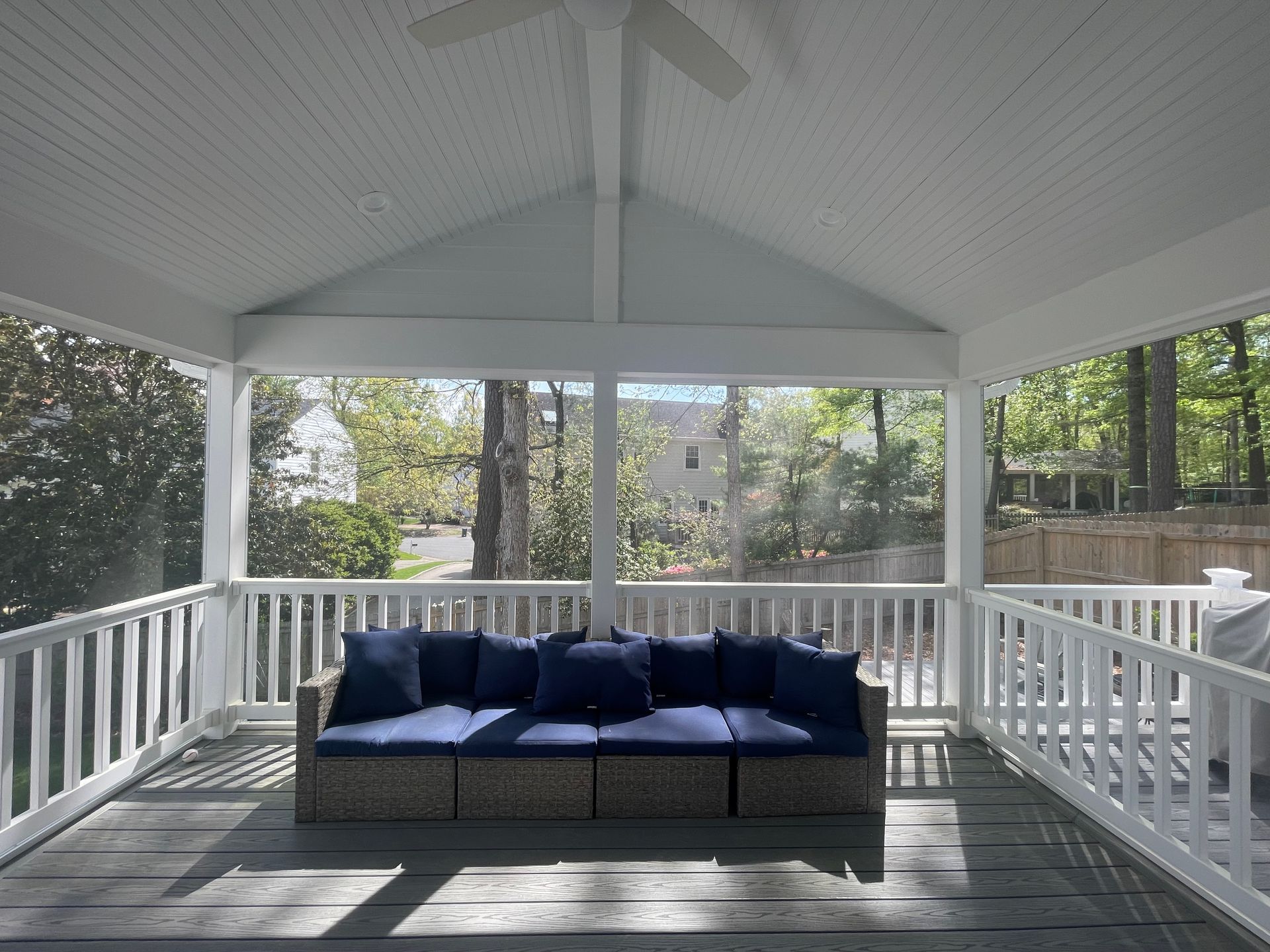 Covered porch with white railings, ceiling, and blue sofa with cushions. Screened windows, view of trees.