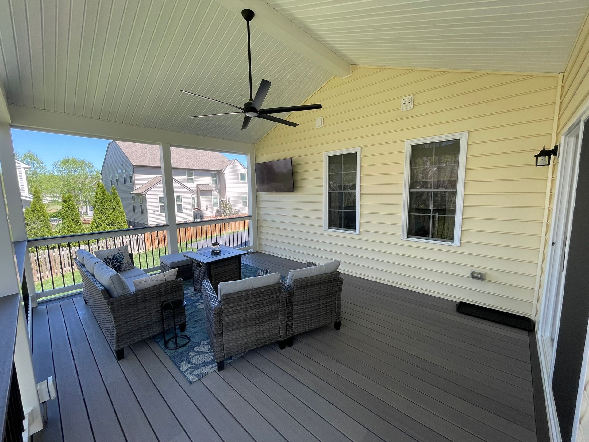 Covered outdoor deck with seating, TV, and ceiling fan; yellow siding, gray deck, view of houses.