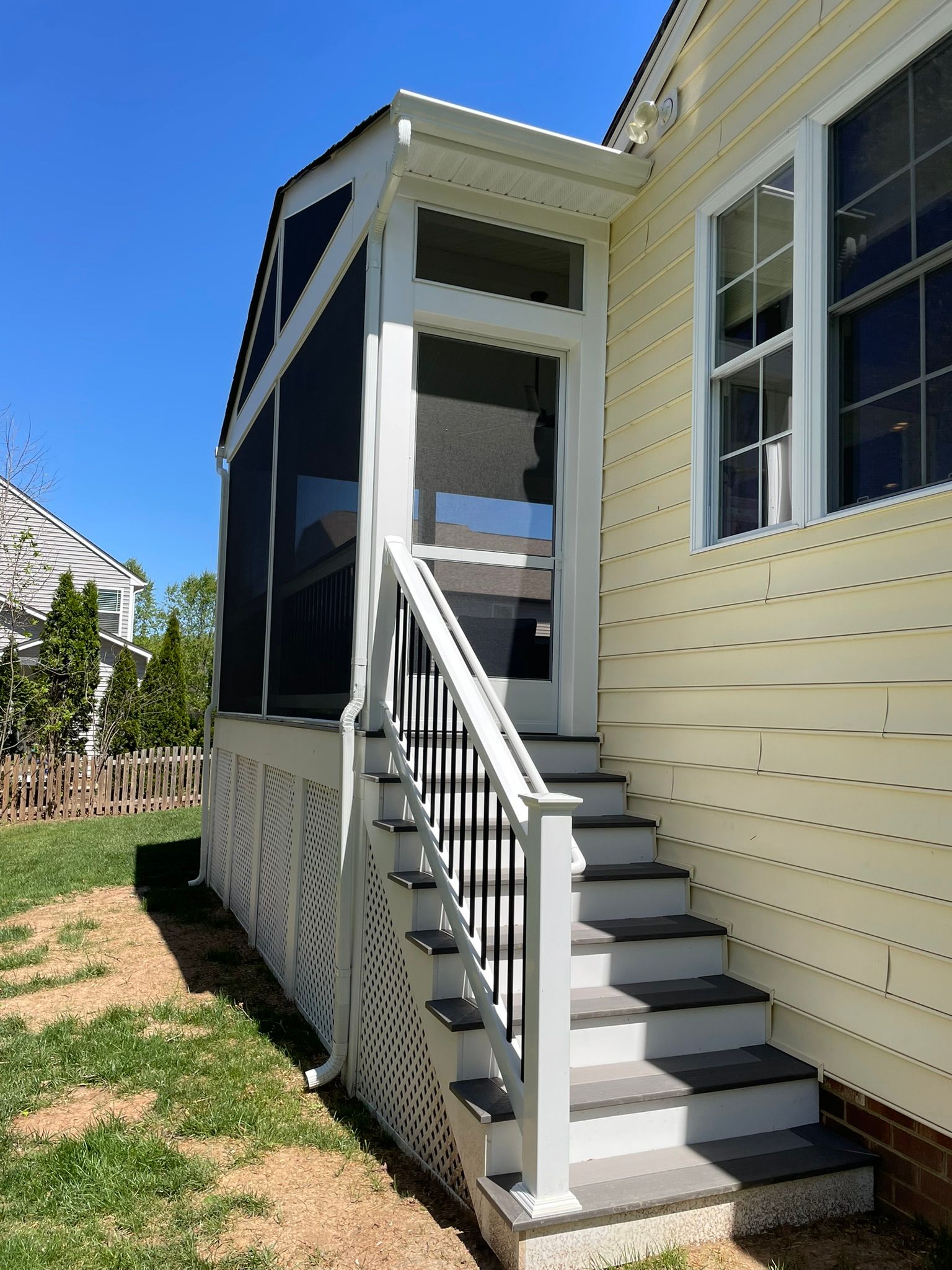 Screened porch with steps and black railing next to a yellow house, on a sunny day.