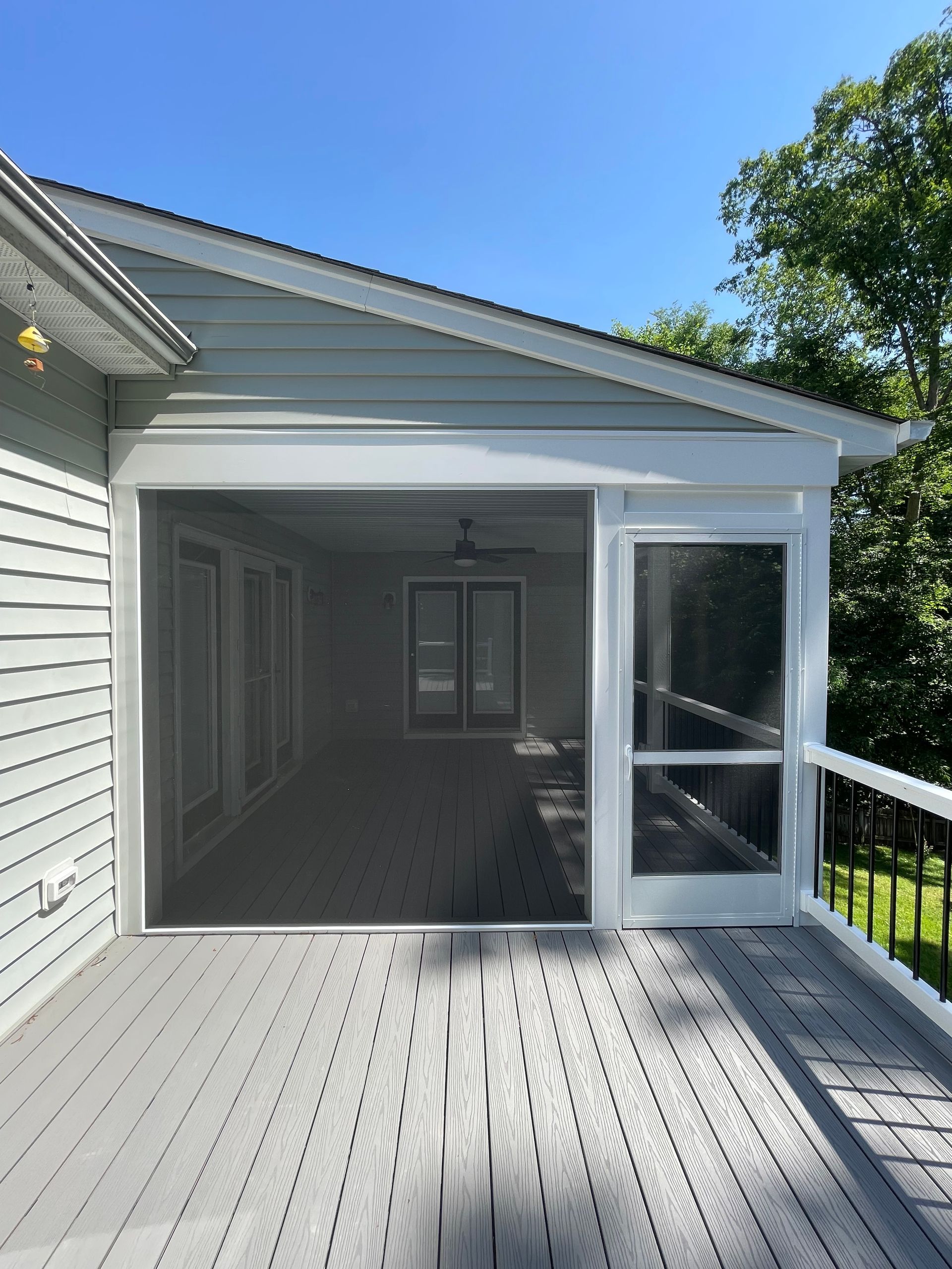 Screened-in porch with gray decking and a white frame, overlooking a green yard under a blue sky.