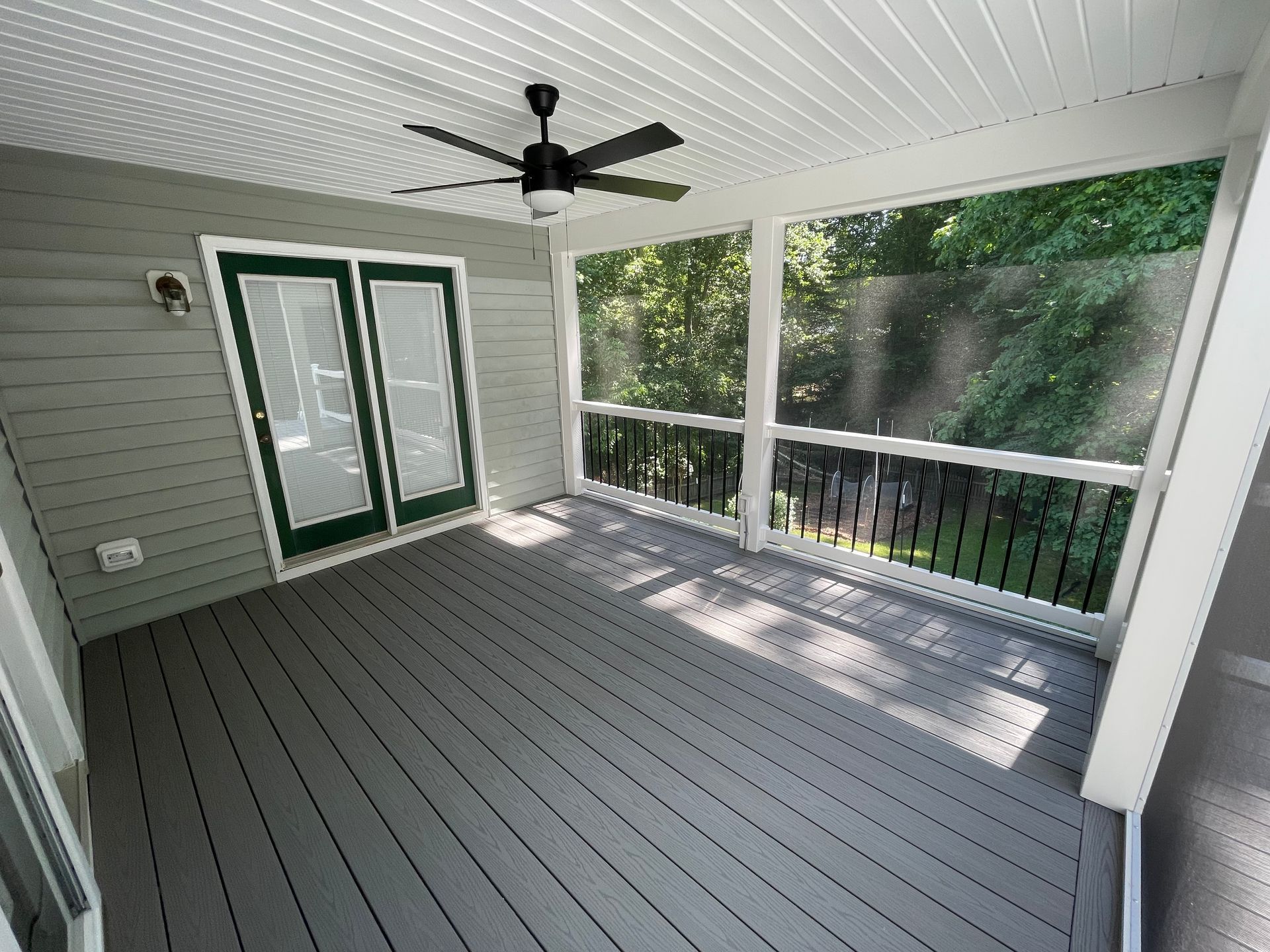 Screened-in porch with gray floor and ceiling fan. Double doors on left, screens and railing on right overlooking trees.