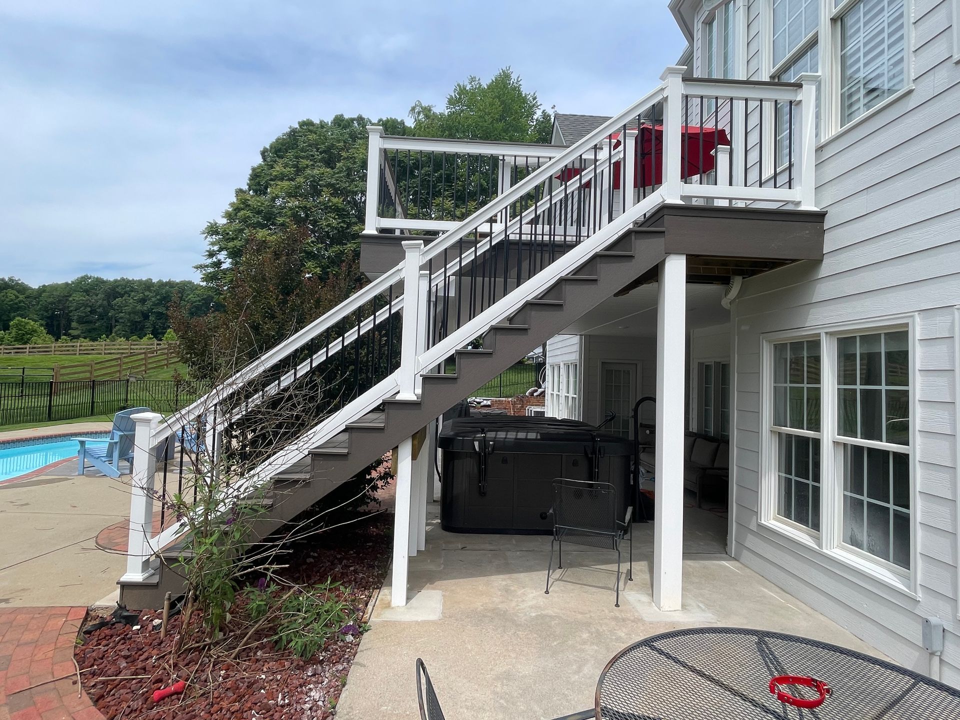 Deck with stairs leading down to a patio and hot tub, attached to a white house with windows.