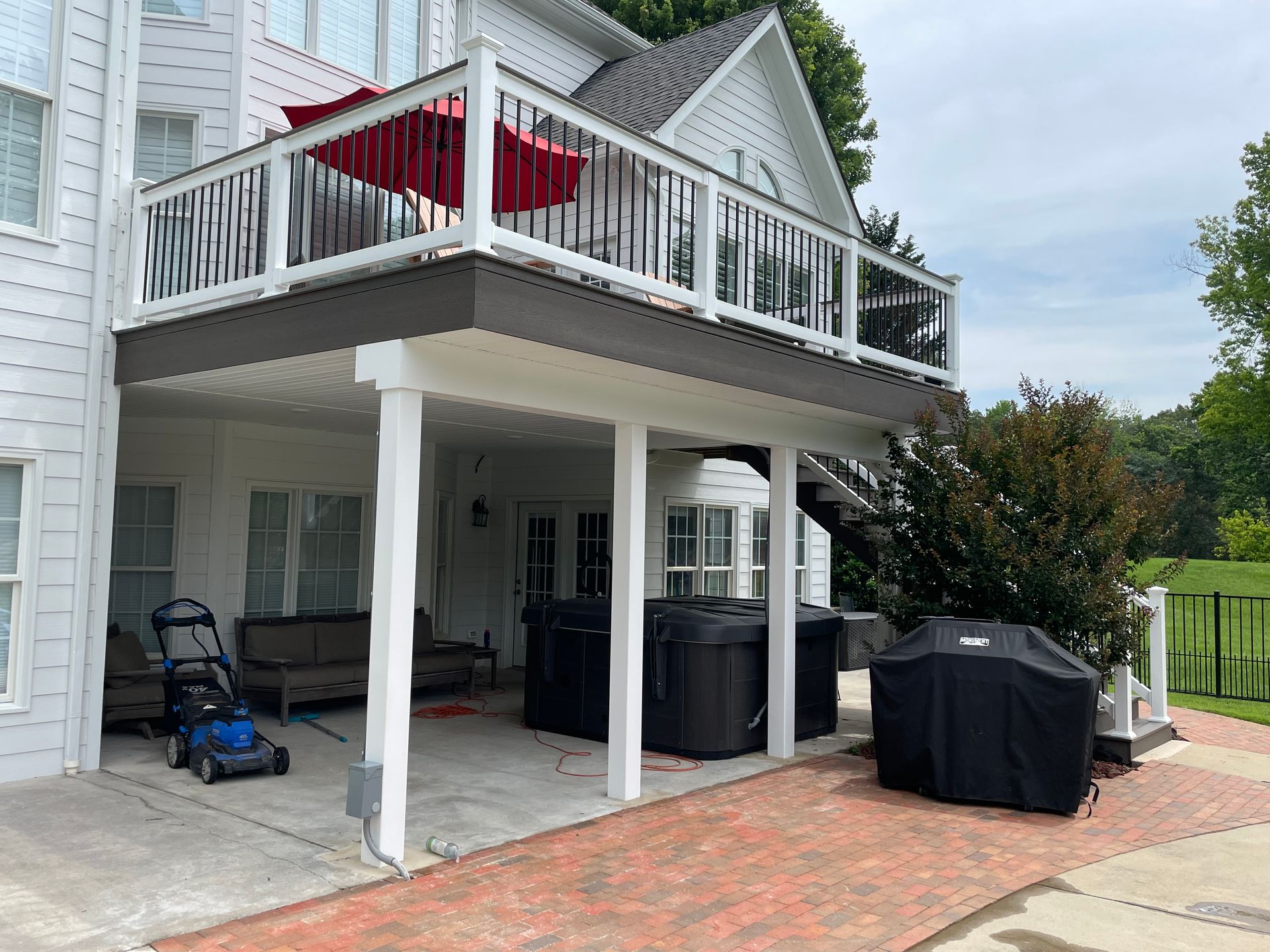 Two-story white house with deck; red umbrella on top deck. Lower level has covered patio with grill, hot tub, and lawnmower.