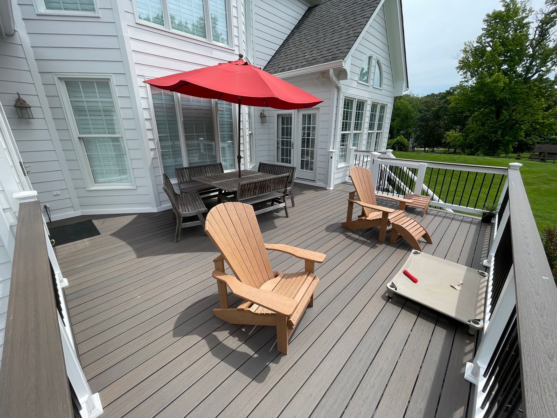 Deck with outdoor furniture: red umbrella, wooden chairs, table, and a dog bed next to a white house.