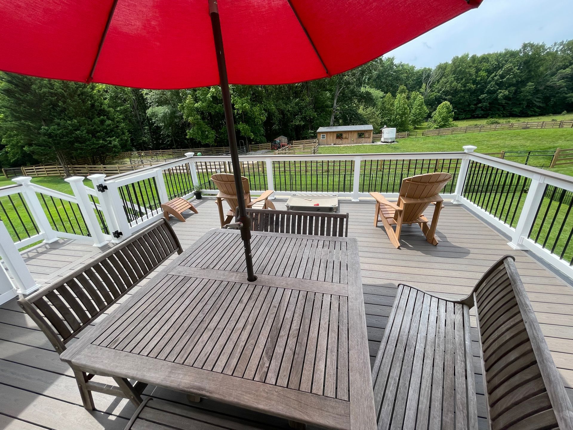 Wooden deck with outdoor furniture under a red umbrella, overlooking a grassy area and trees.