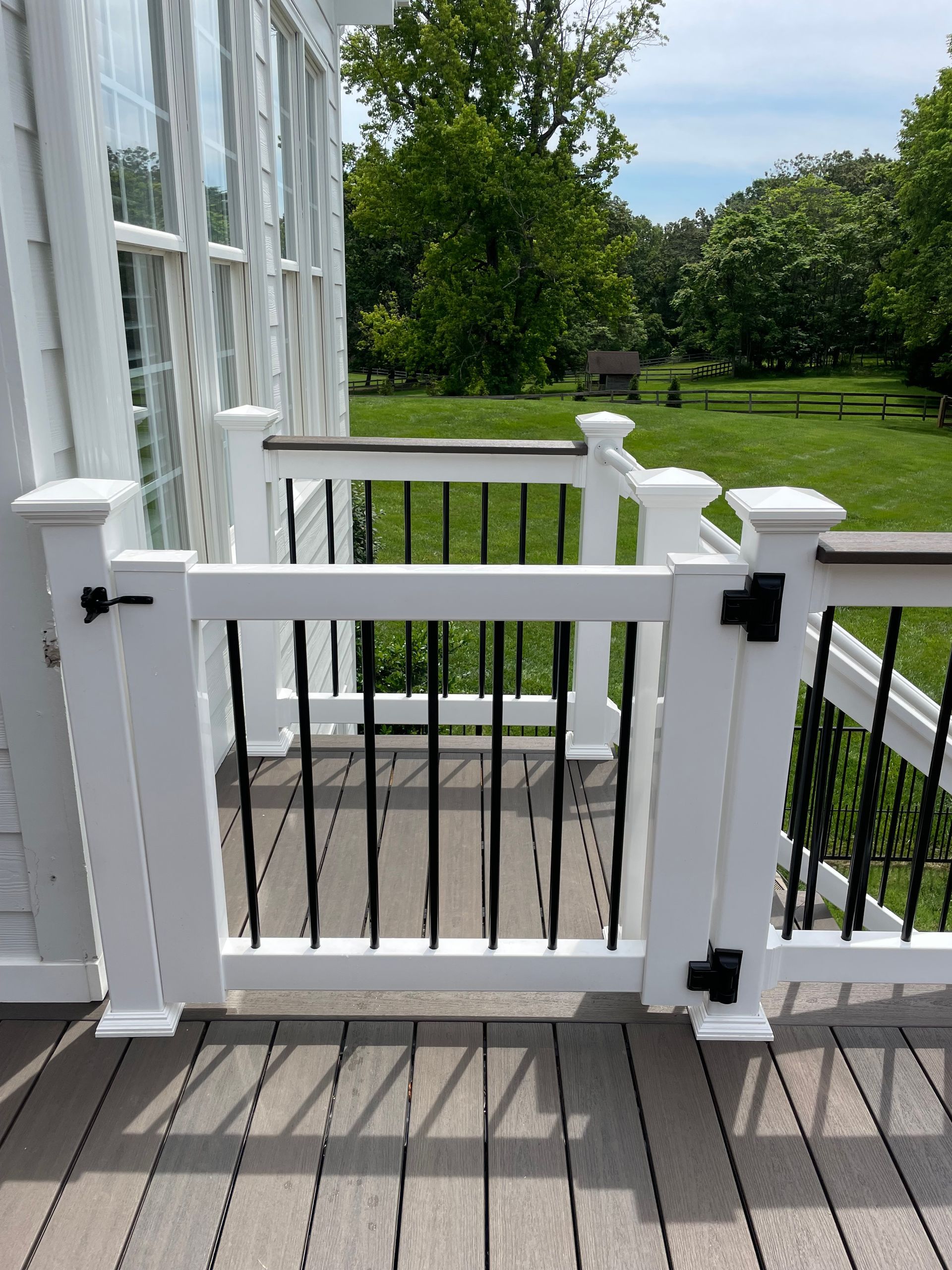 White picket gate with black bars, attached to a wooden deck, overlooking a green lawn.