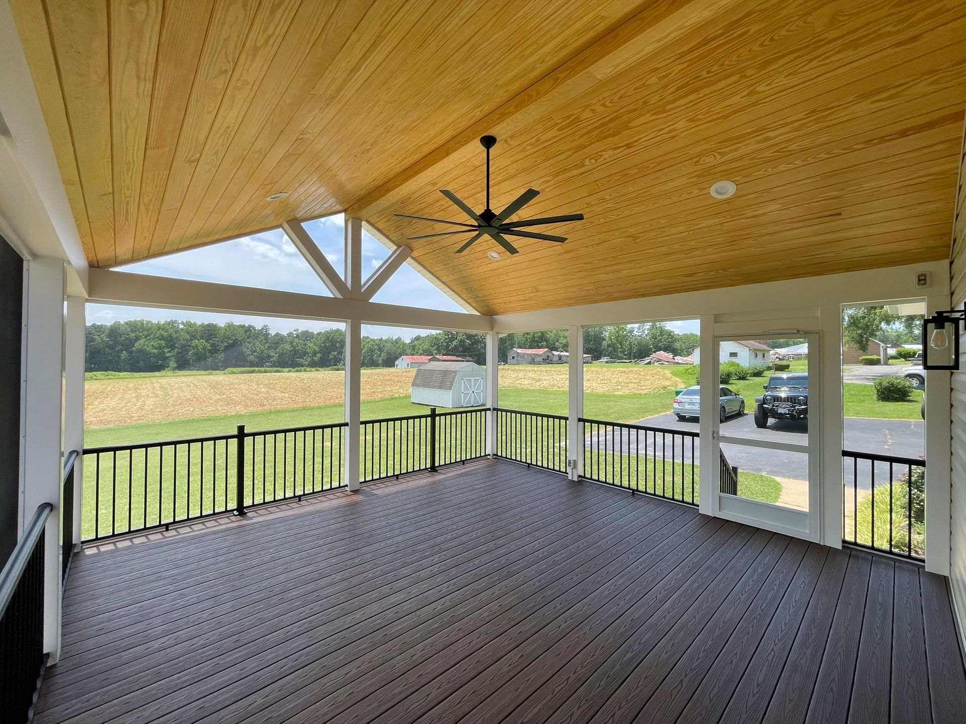 Covered porch with wood ceiling and brown deck overlooking a grassy field. Black railing and ceiling fan.