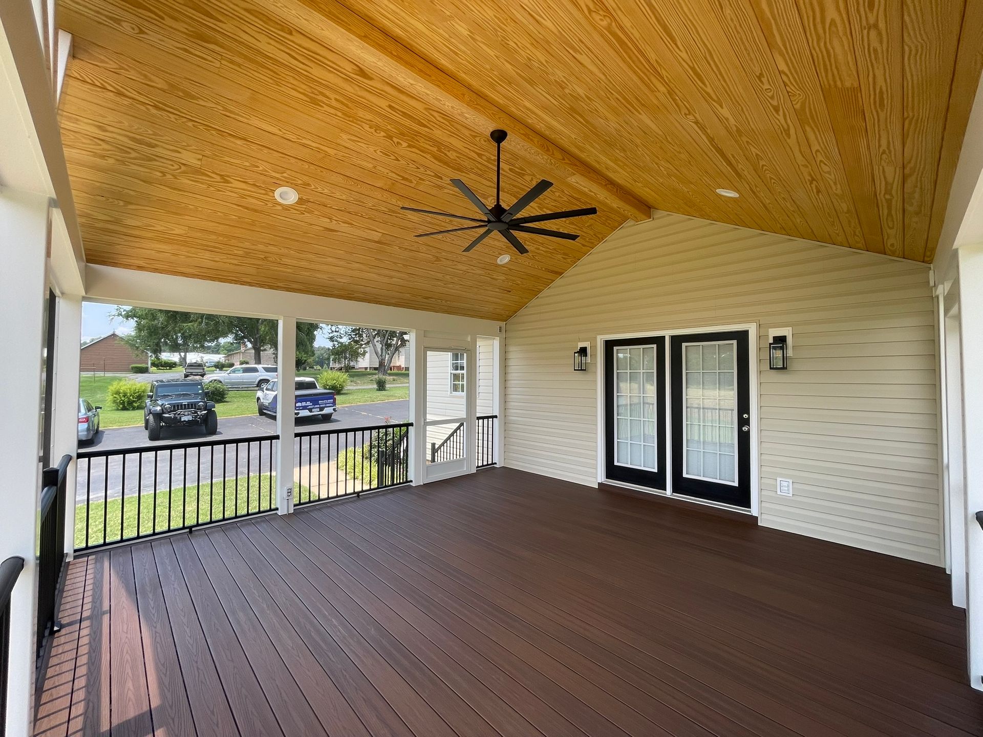 Covered porch with wood ceiling, dark composite decking, black railing, and double doors.
