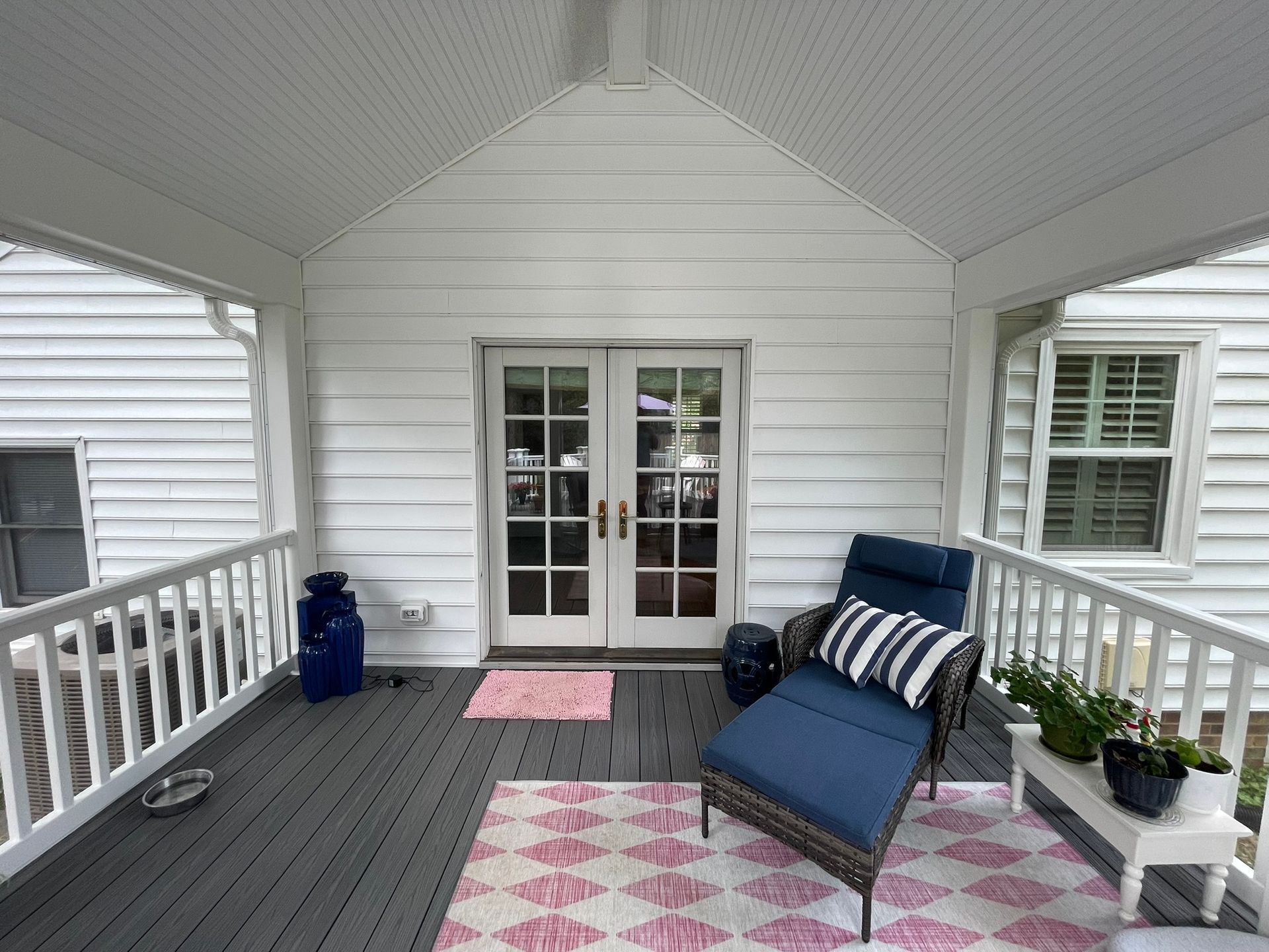 Covered porch with white railings, gray floor, blue chaise, and pink rugs.