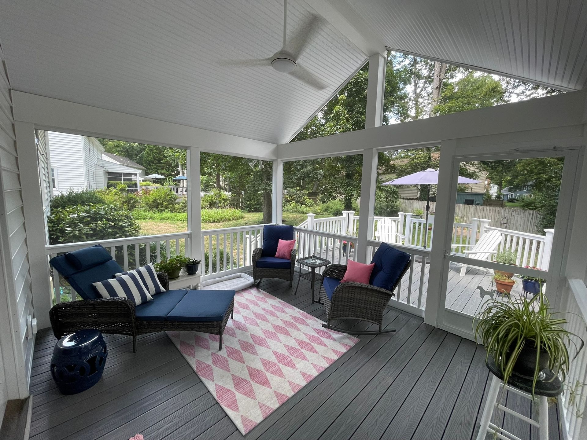Screened-in porch with blue furniture, pink rug, and a ceiling fan; overlooking a backyard.