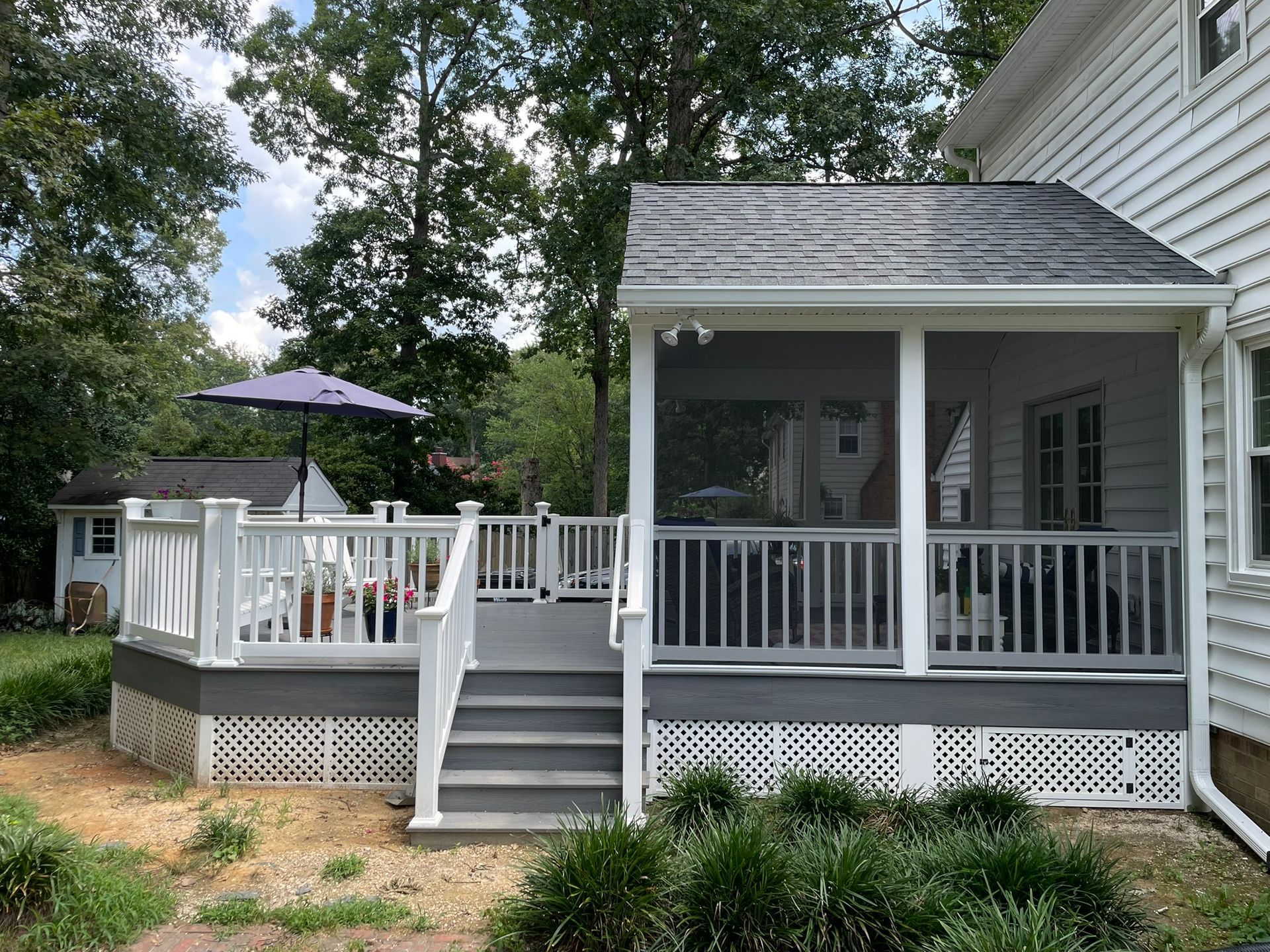 Screened porch and deck attached to a house with gray and white features, surrounded by greenery.