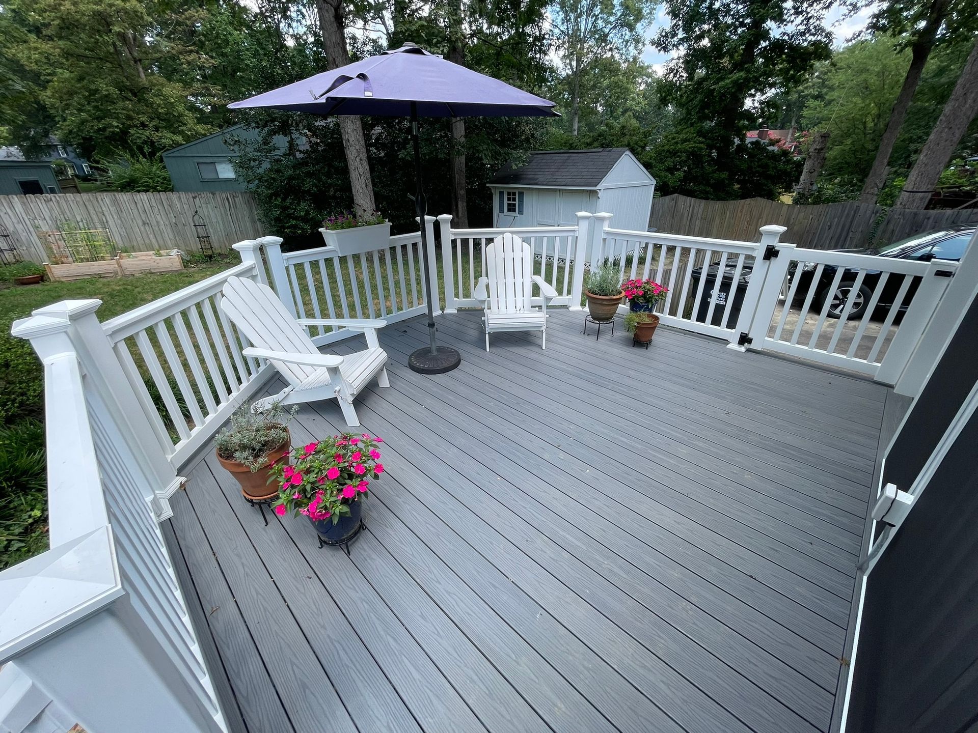 A gray composite deck with white railing, Adirondack chairs, an umbrella, and potted flowers.