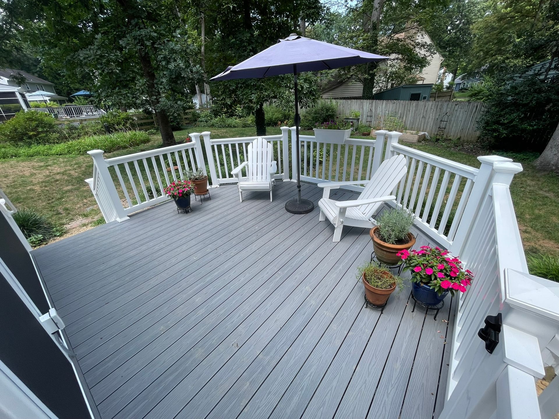Gray composite deck with white railing, two white chairs, purple umbrella, and potted flowers.
