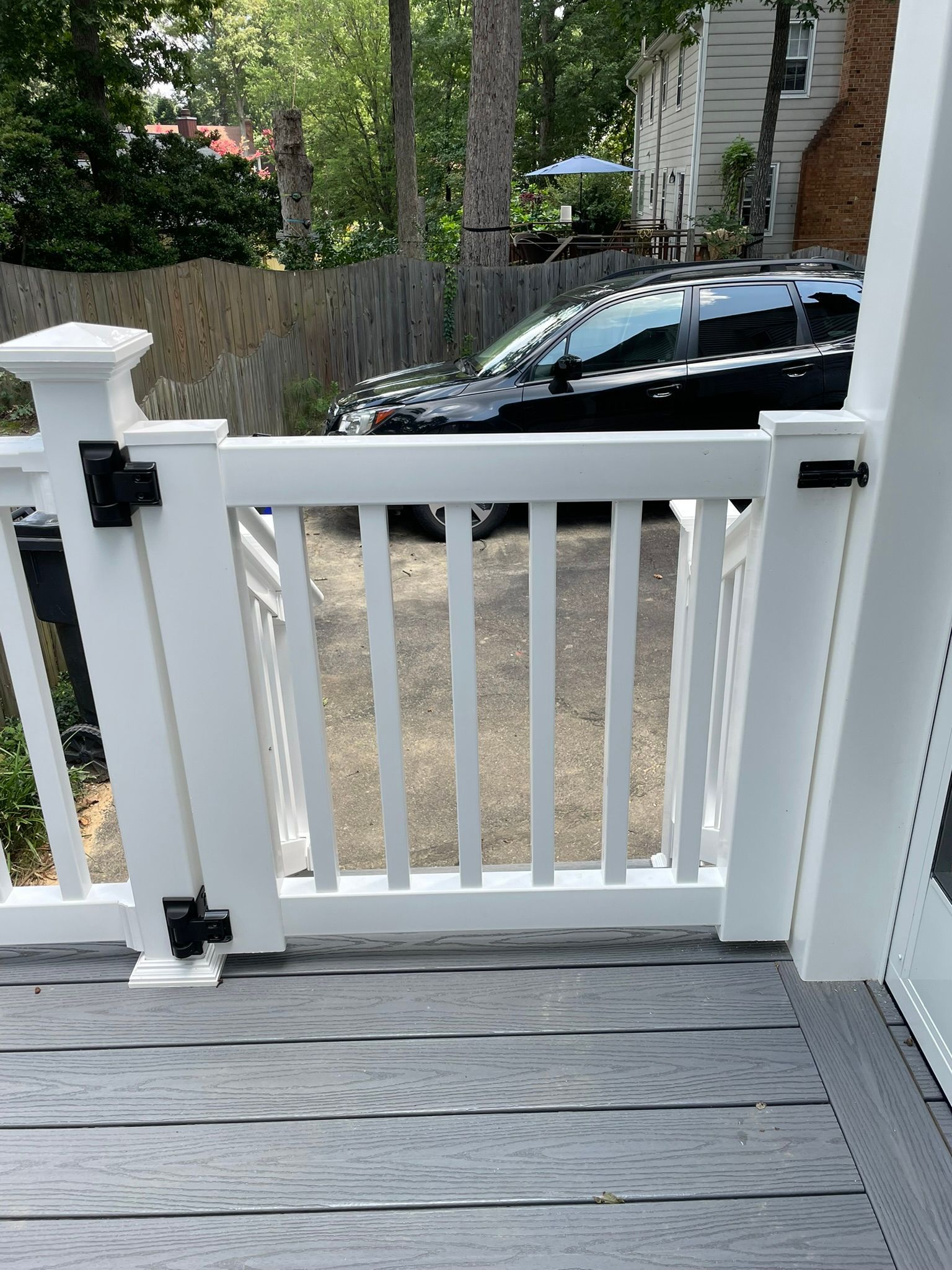 White picket gate on a gray deck, with black hinges, leading to a gravel driveway with a black SUV parked.