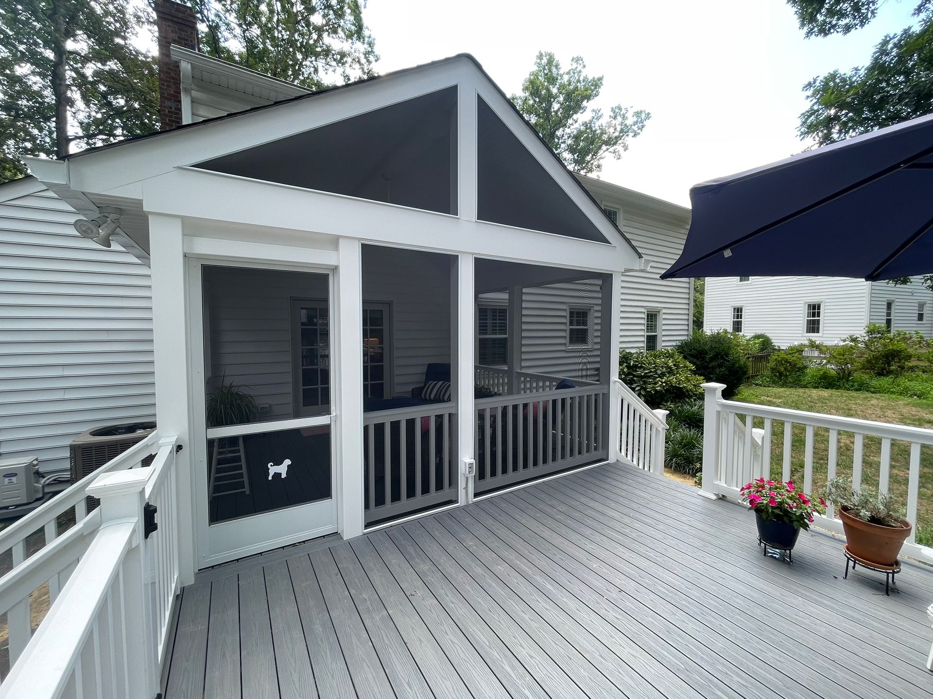 Screened porch with gray deck and white railing, attached to a house. A blue umbrella is partially visible.