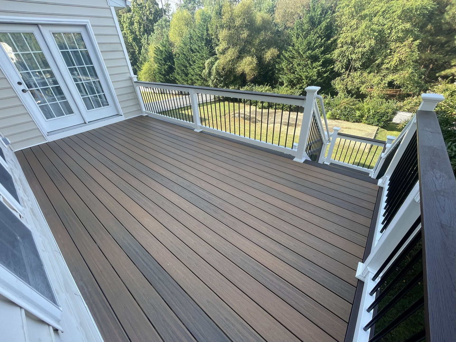Composite deck with black railing and white posts, next to a house with French doors; trees in the background.