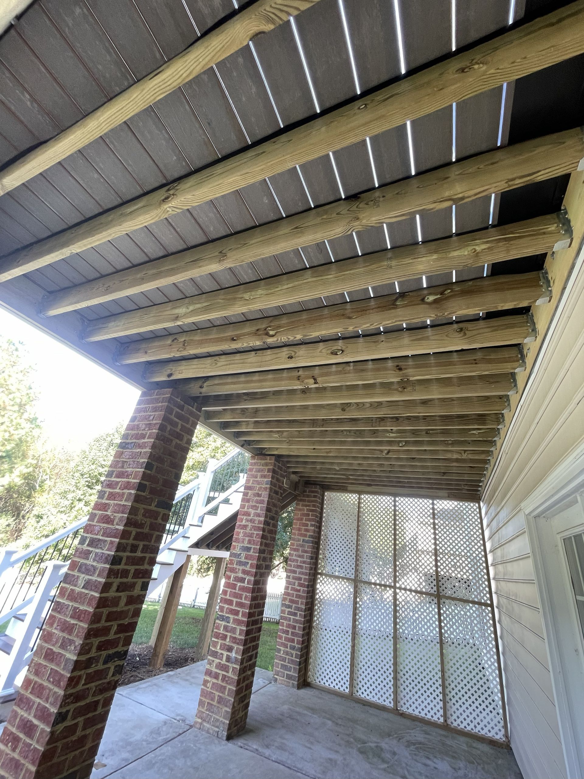 Underside view of a wooden deck supported by brick pillars, with a concrete patio below.