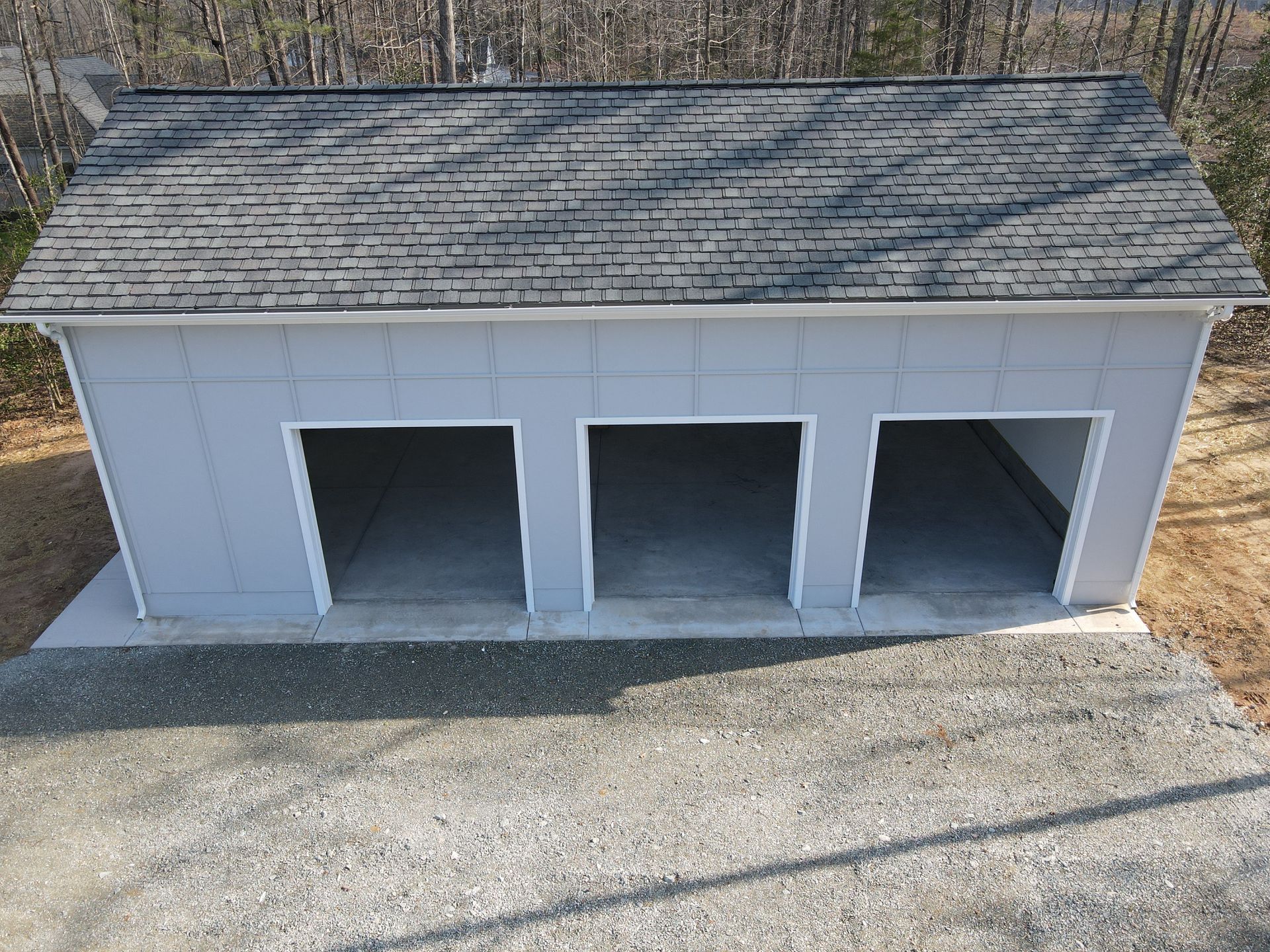 Three-bay, gray garage with open bays. Gravel driveway in foreground. Roof is gray asphalt shingles.