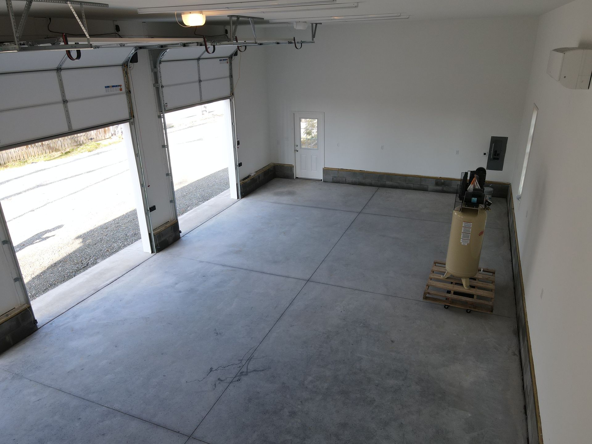 Empty garage with open doors, concrete floor, white walls, compressor on a pallet.
