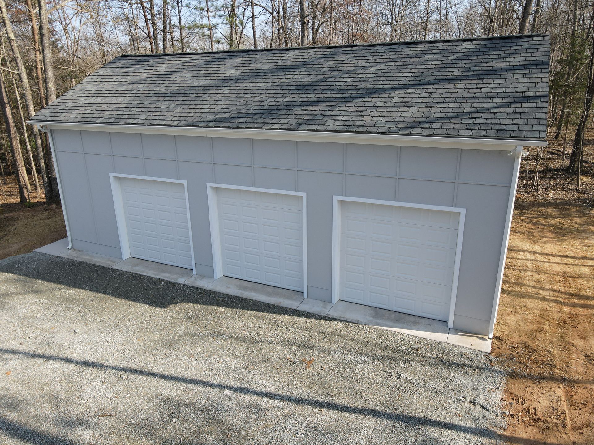 Three-car garage with gray siding, white doors, and a gray shingled roof on a gravel driveway.