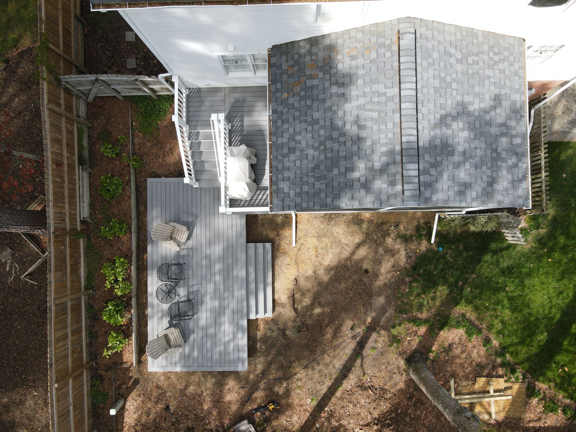 Aerial view of a house with a gray shingle roof, a deck, and a yard with trees and a fence.