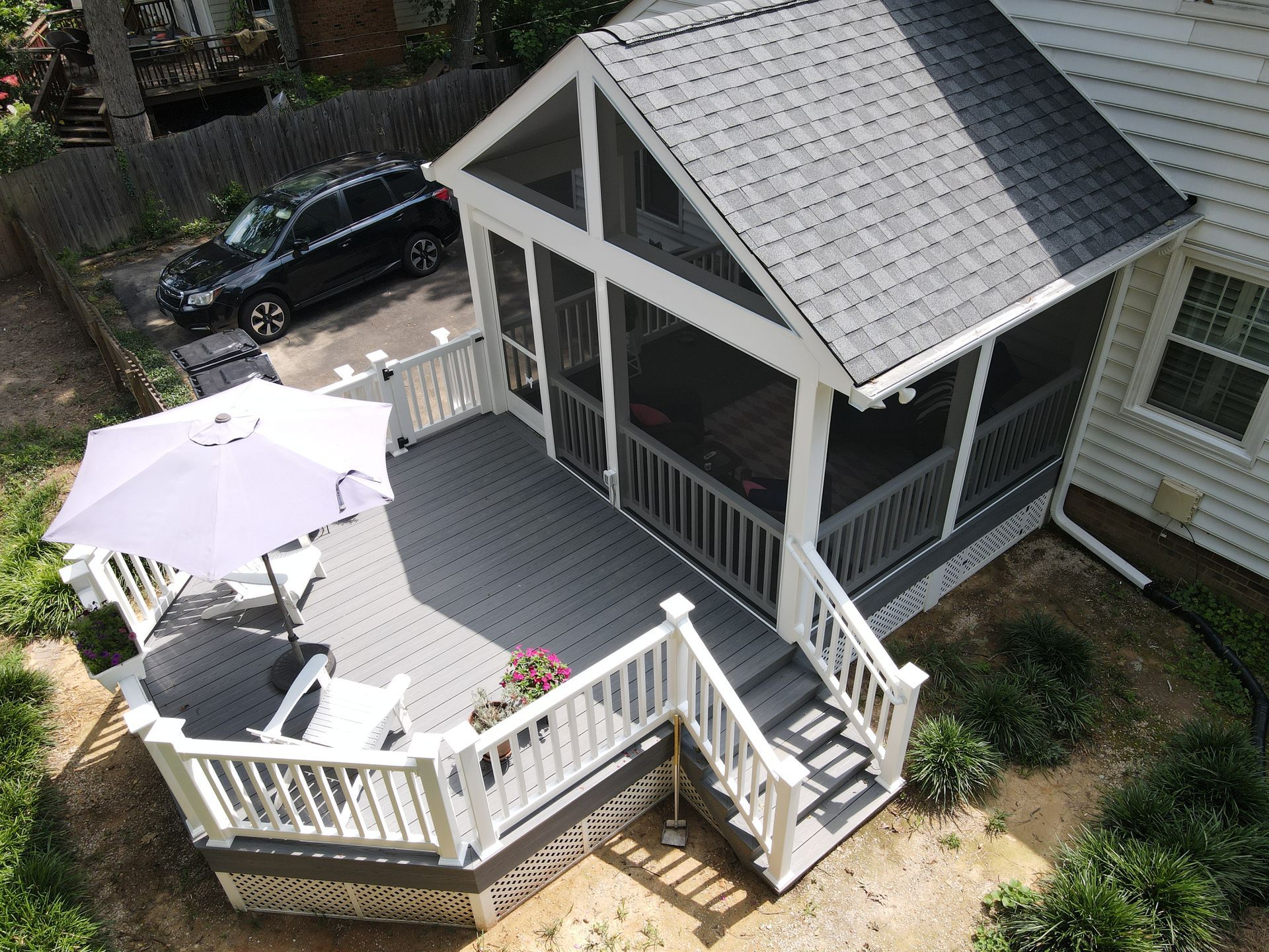 Screened porch with gray deck, white railings, and a gray roof, next to a home and a car.