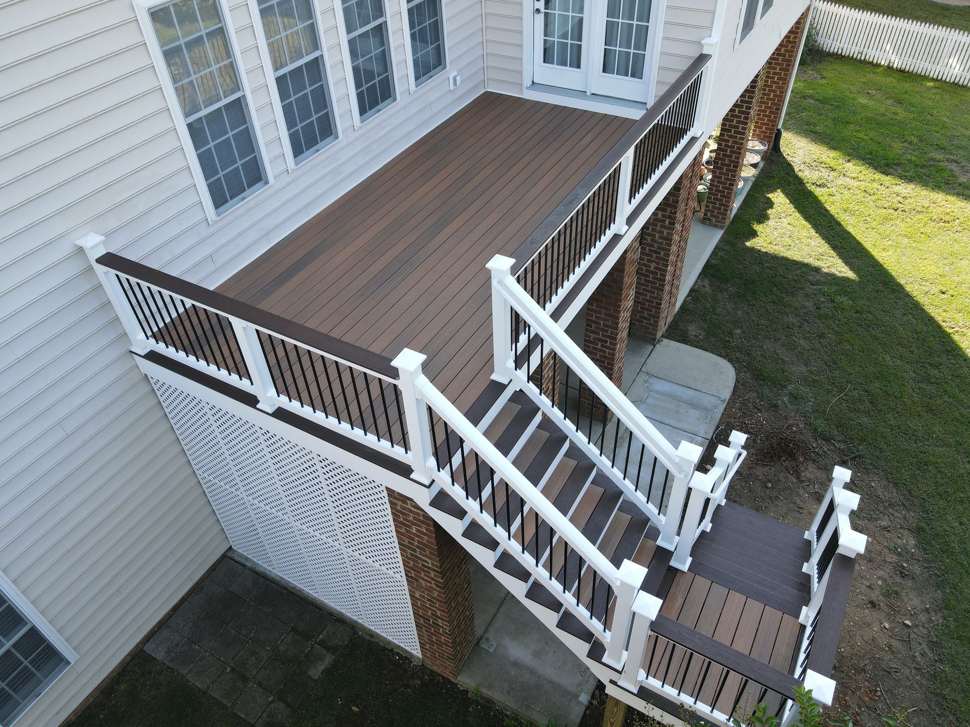 Elevated deck with brown composite boards, white railing and stairs against a beige house with brick supports.