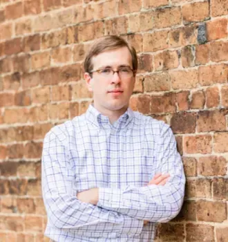 A man in a plaid shirt is standing in front of a brick wall with his arms crossed.