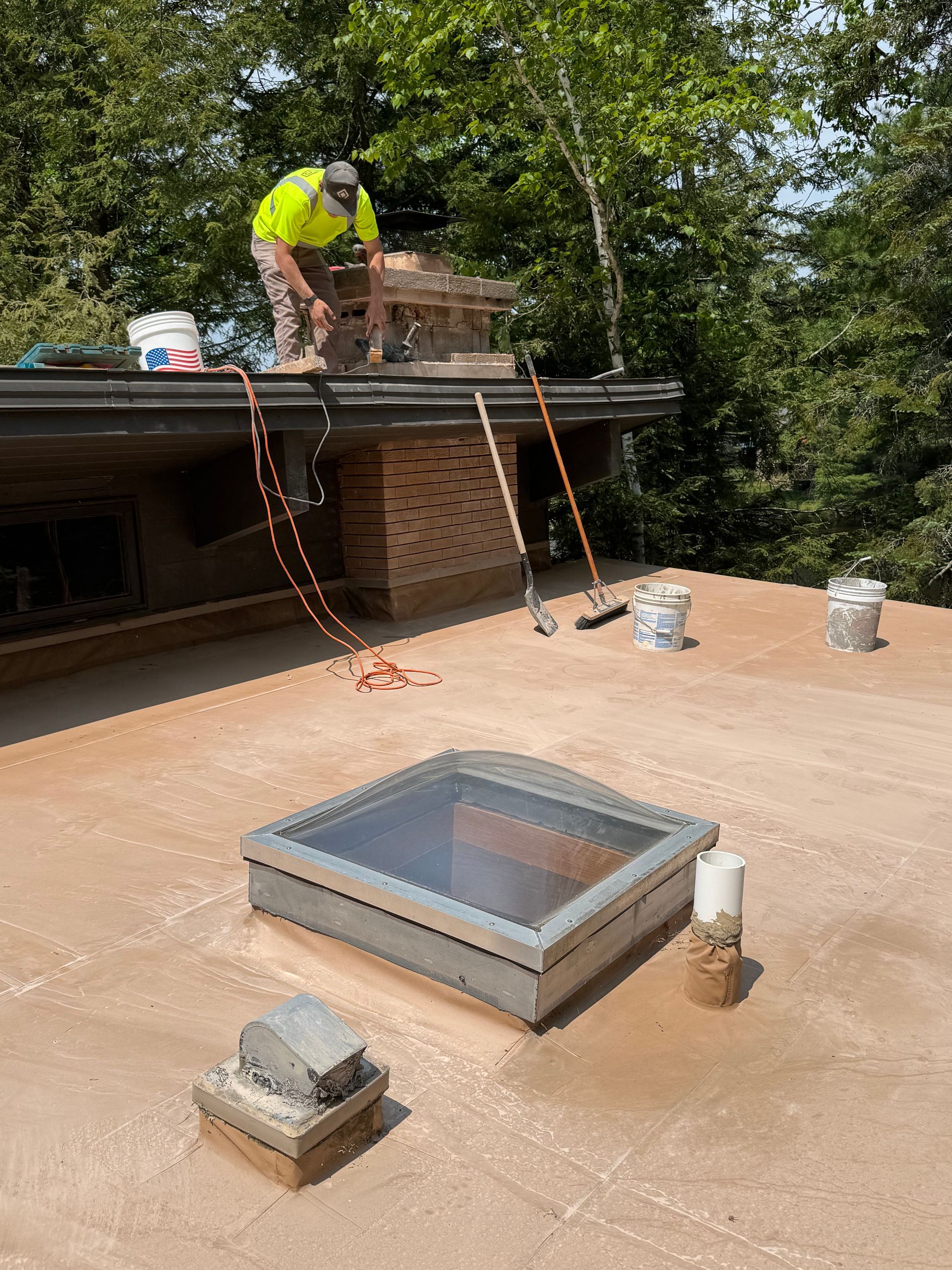 Person on a roof repairing a brick chimney; flat roof with skylight, tools, and buckets.