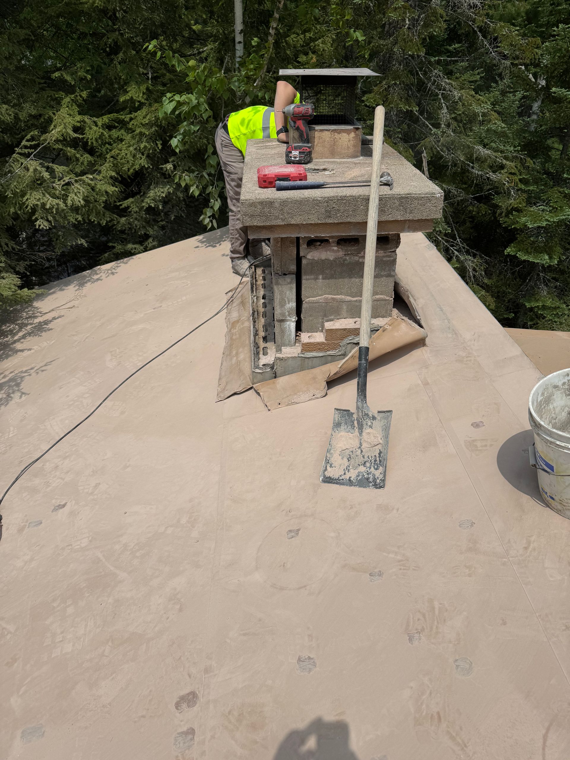 Roofer repairing chimney on a rooftop. A shovel, bucket, and tools are nearby. Green trees in the background.