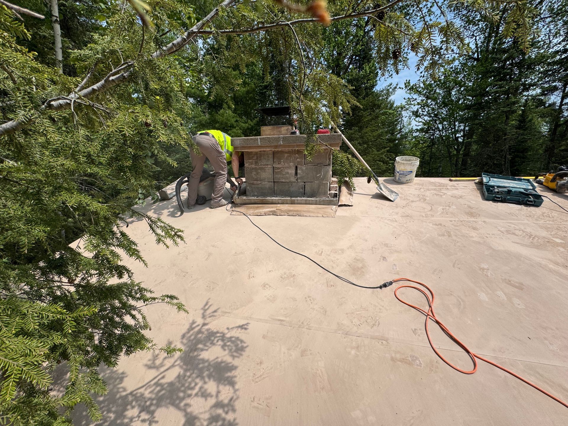A person using a saw to work on a brick chimney on a flat rooftop, under a sunny sky.