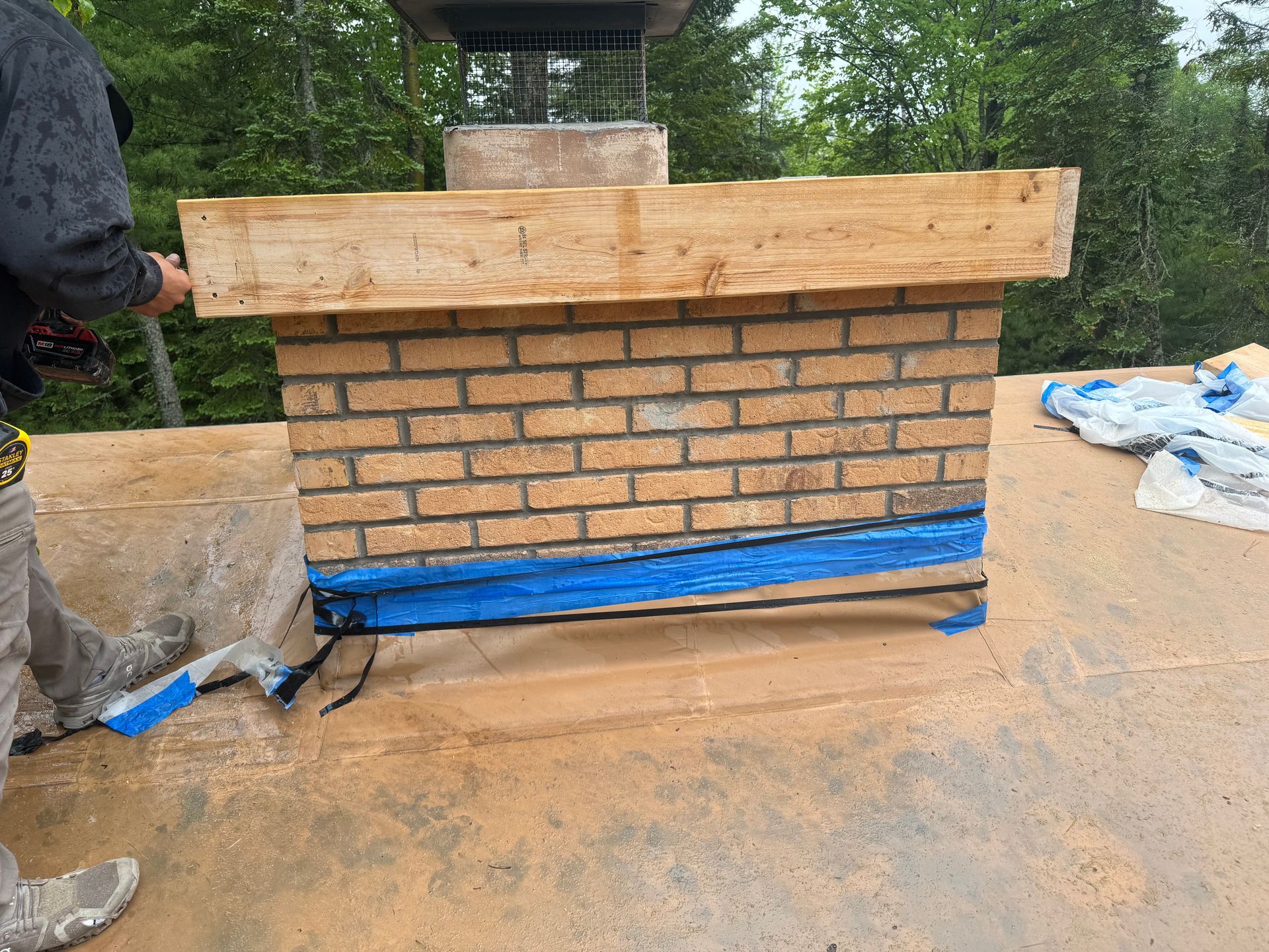 Man working on a brick chimney on a roof. Blue tape and wood framing are visible.