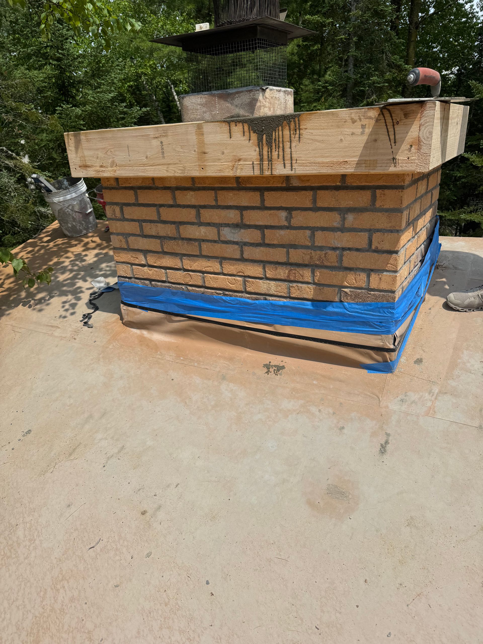 Brick chimney on a flat roof, blue flashing tape, surrounding tree background, and a metal chimney cap.
